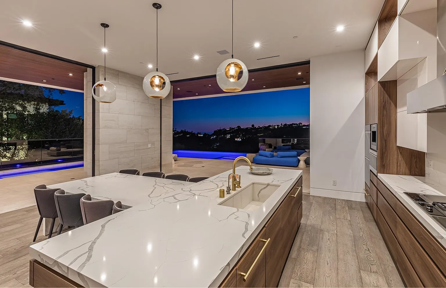 Kitchen island at nightfall, glass pendants lit and the pool terrace and hillside lights framed by open walls
