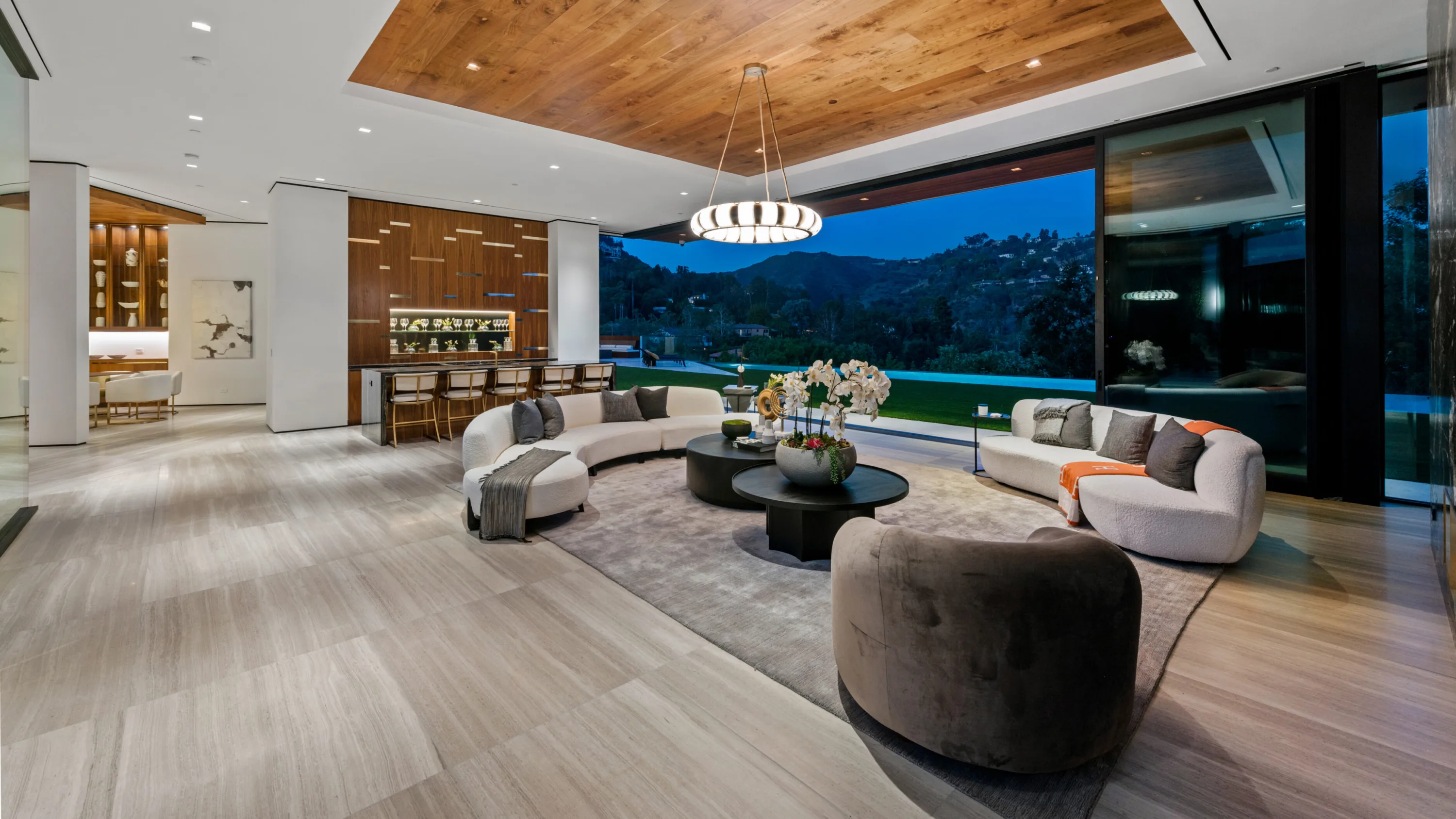 Same lounge and bar after dark, drum chandelier glowing under the timber ceiling as the hills fade