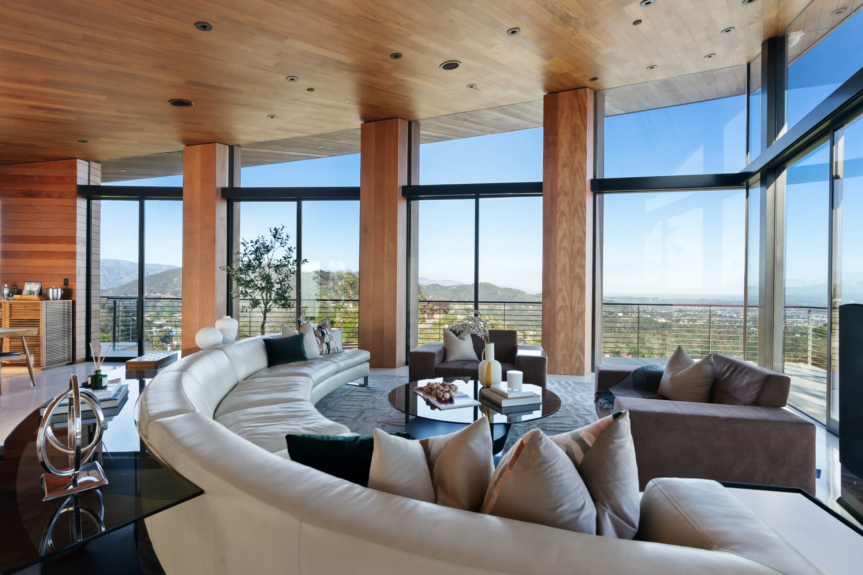 Curved cream sectional in the glass-walled living room, timber ceiling above and hillside city views on two sides