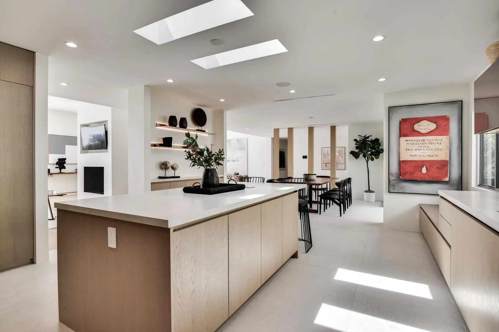 Long pale oak island under twin skylights, open to the dining table and lounge beyond