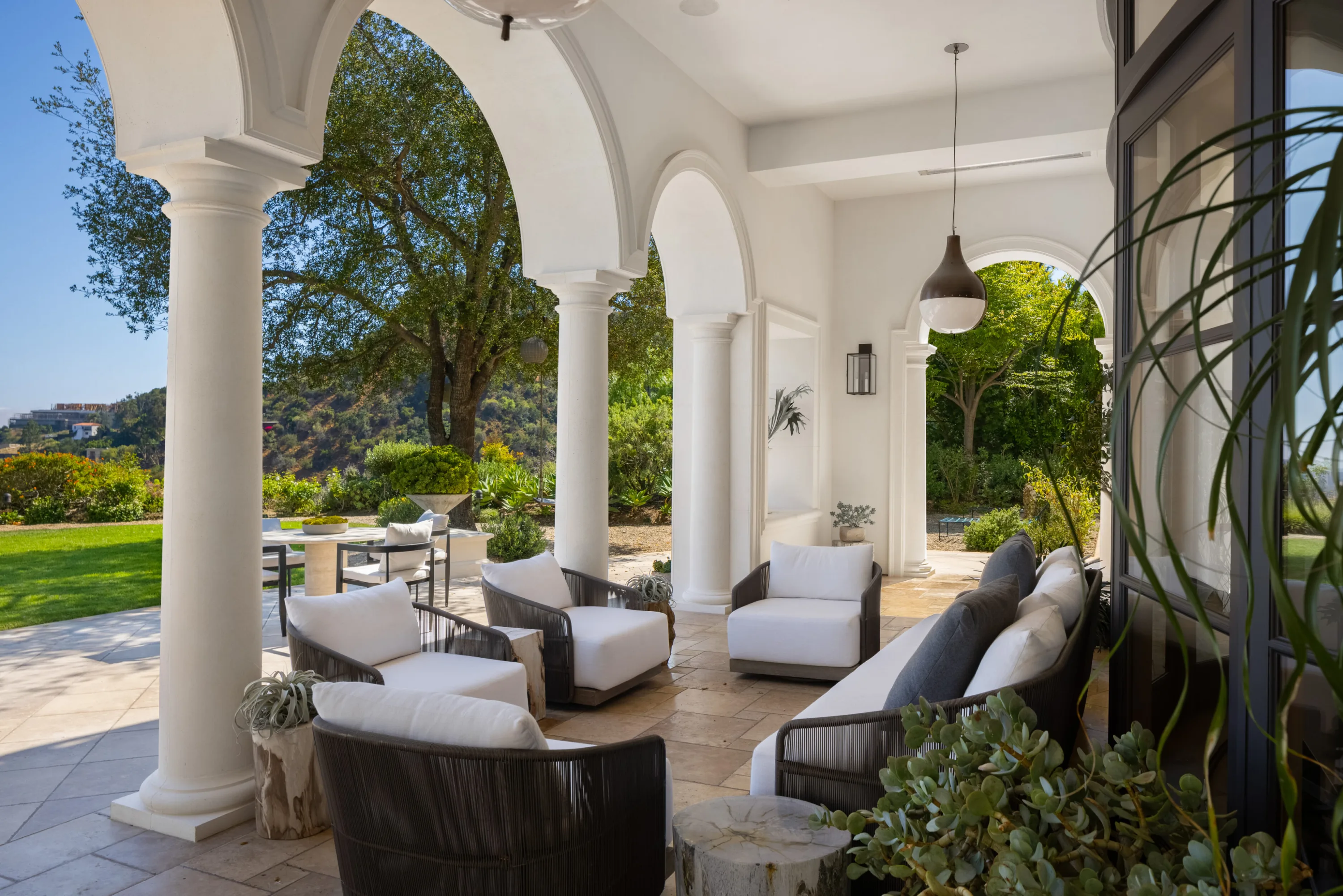 Arched loggia lounge with woven armchairs and a dining table looking out to lawn and hillside