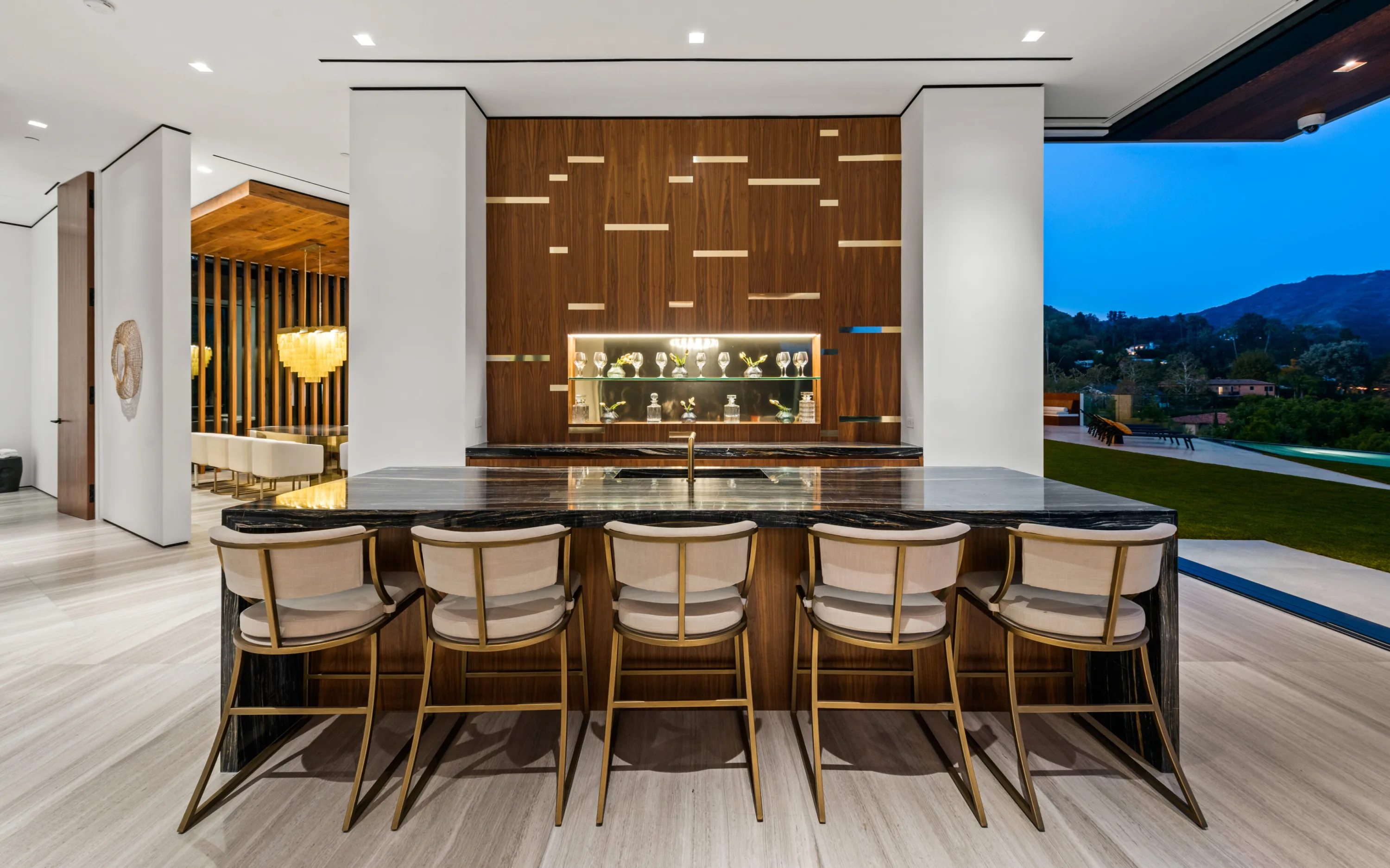 Walnut and dark-marble bar island with five brass-framed stools, backlit glassware shelves behind