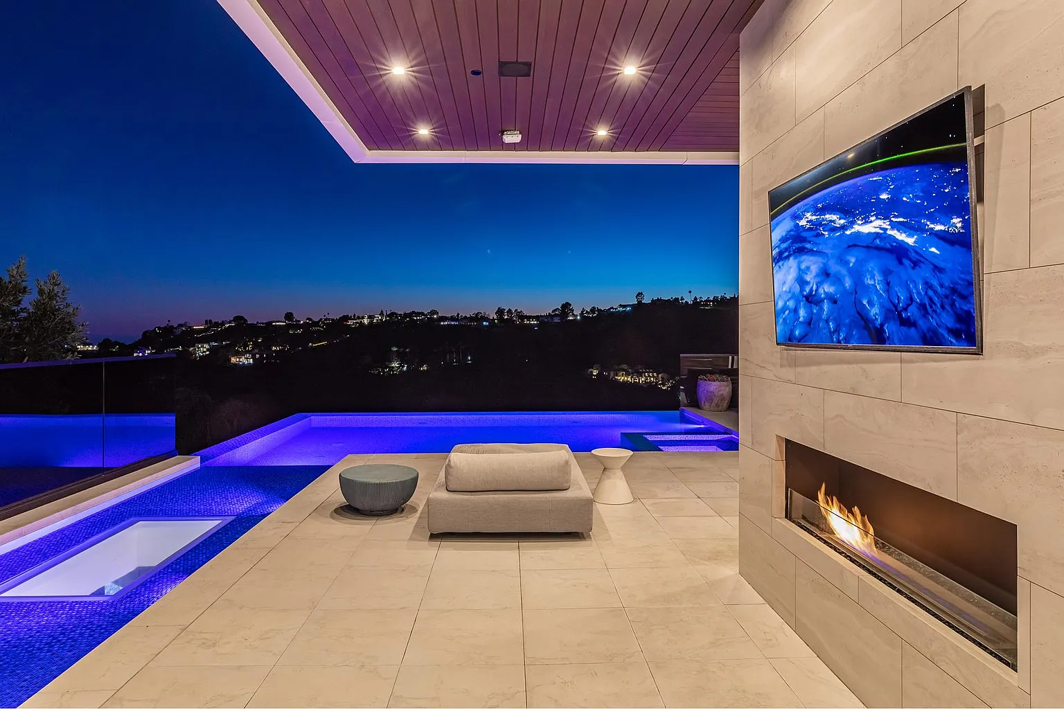 Covered pool deck with a daybed, burning linear fireplace and mounted screen above the illuminated spa