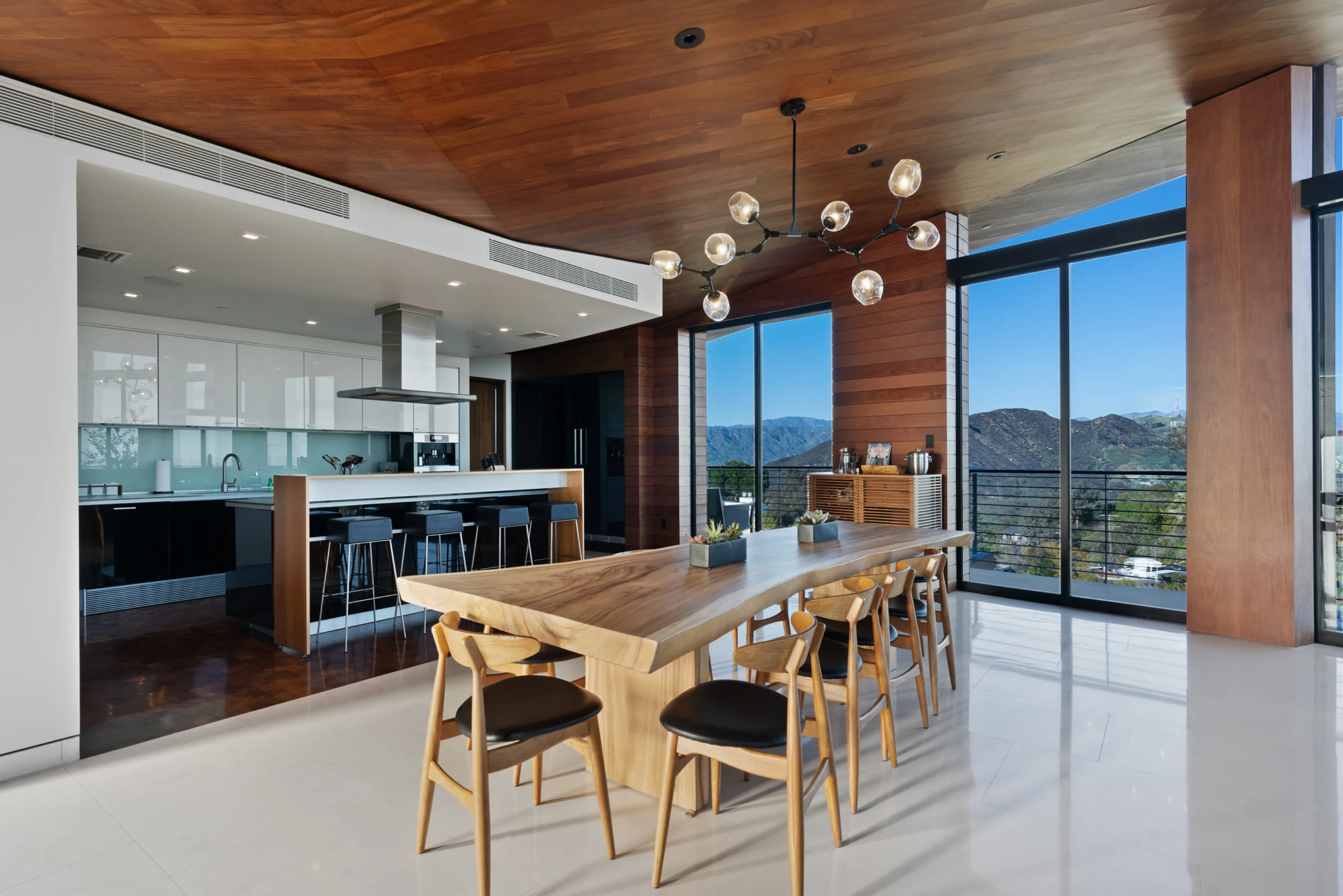 Live-edge dining table for ten under a branching globe chandelier, open to the white kitchen and canyon windows