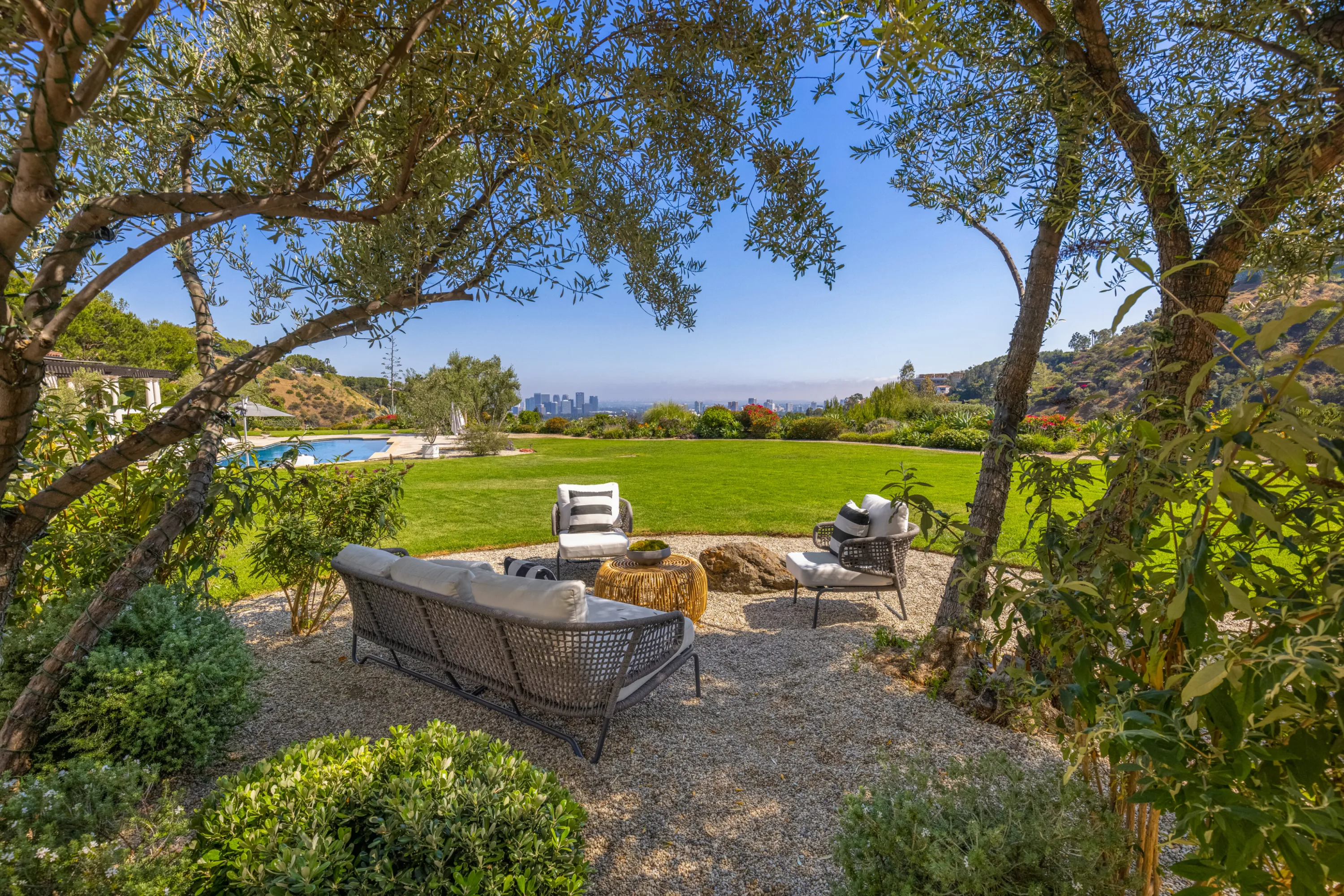 Gravel garden seating circle under olive trees with the lawn, pool and city skyline beyond