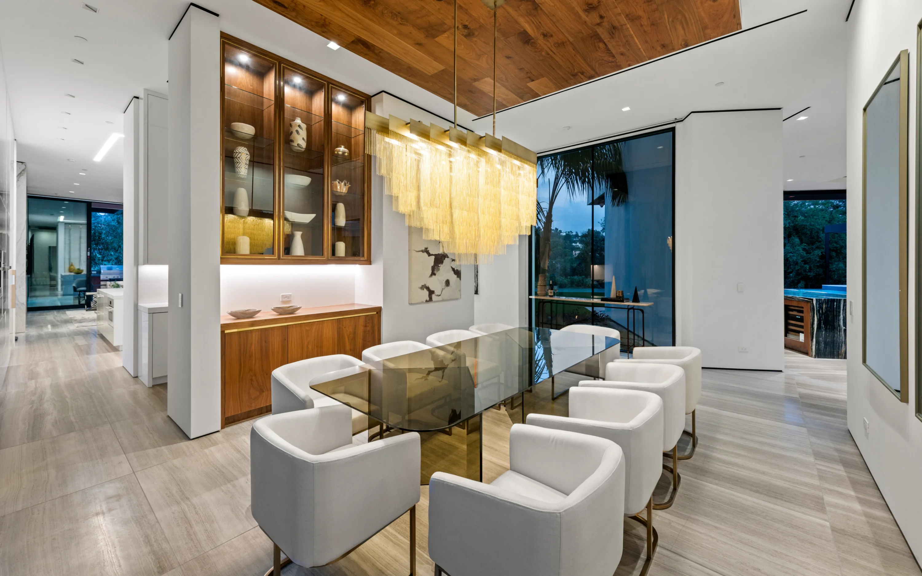Dining area from the hall, glass table and cream armchairs beside a lit walnut display cabinet
