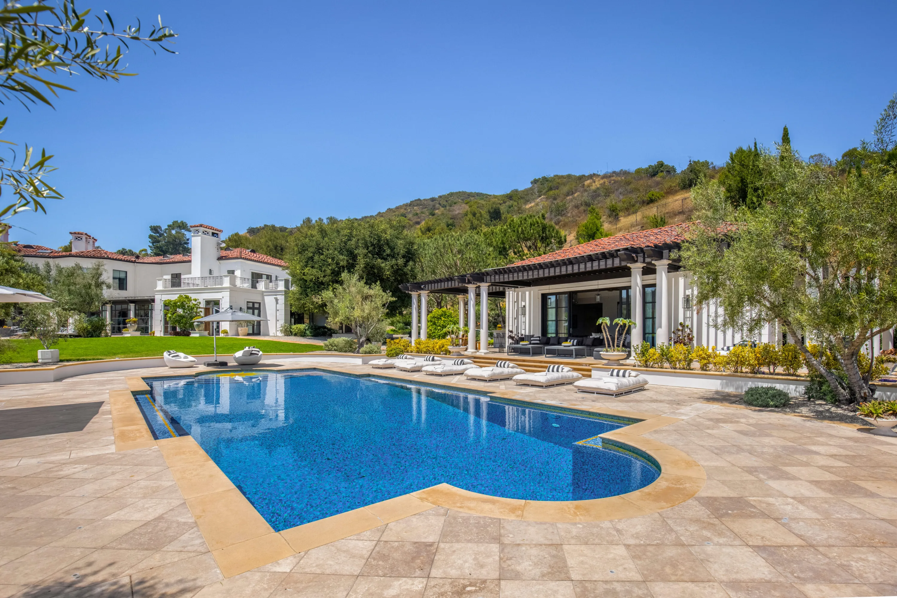 Sun-drenched pool terrace with striped loungers between the main house and the pergola-shaded pool house