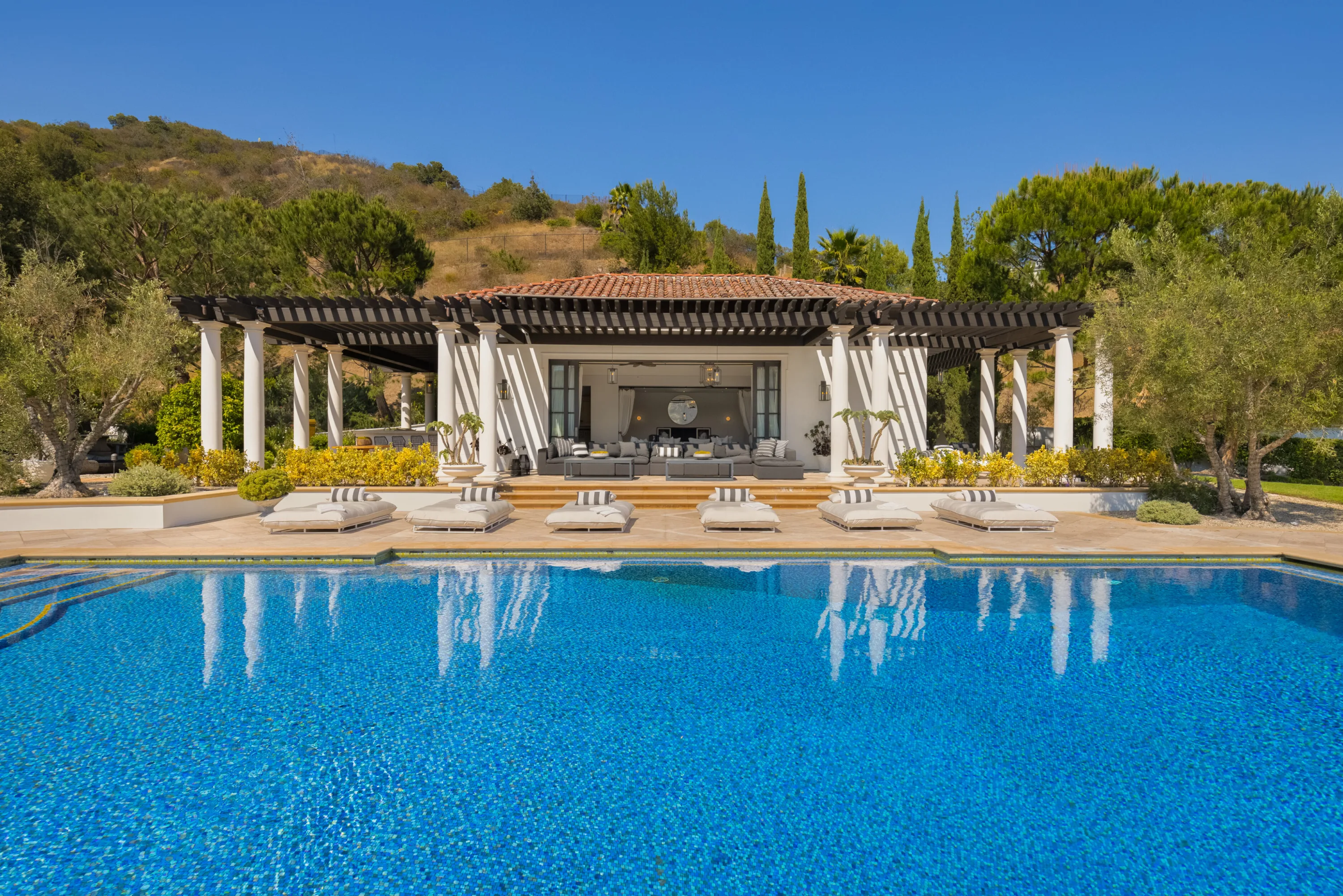 Open-air pool house lounge behind a row of loungers, columns reflected in the blue water