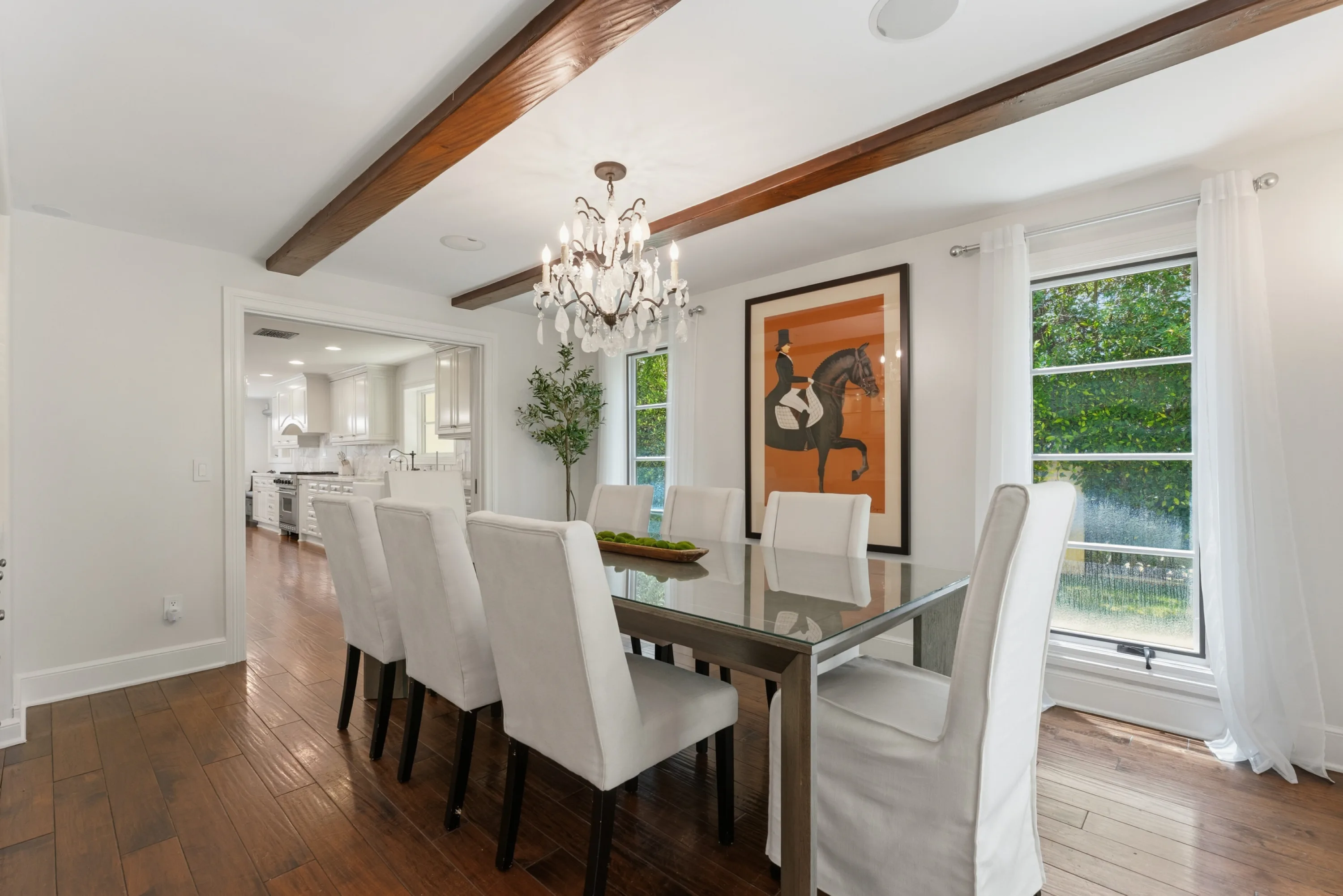 Dining room seating eight beneath a crystal chandelier and exposed beams, open to the kitchen