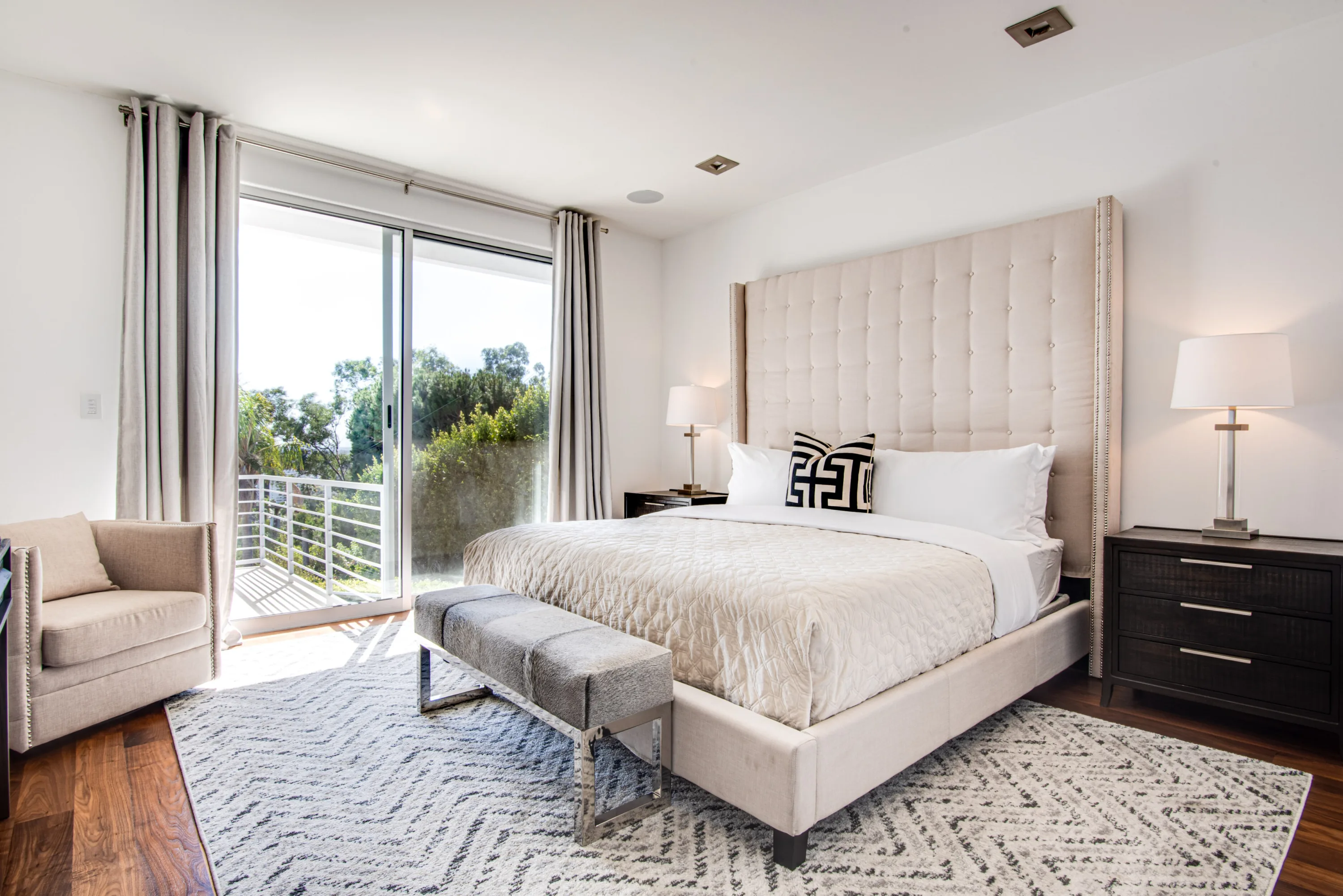 Bedroom with tall tufted headboard and sliding door to a private balcony screened by trees