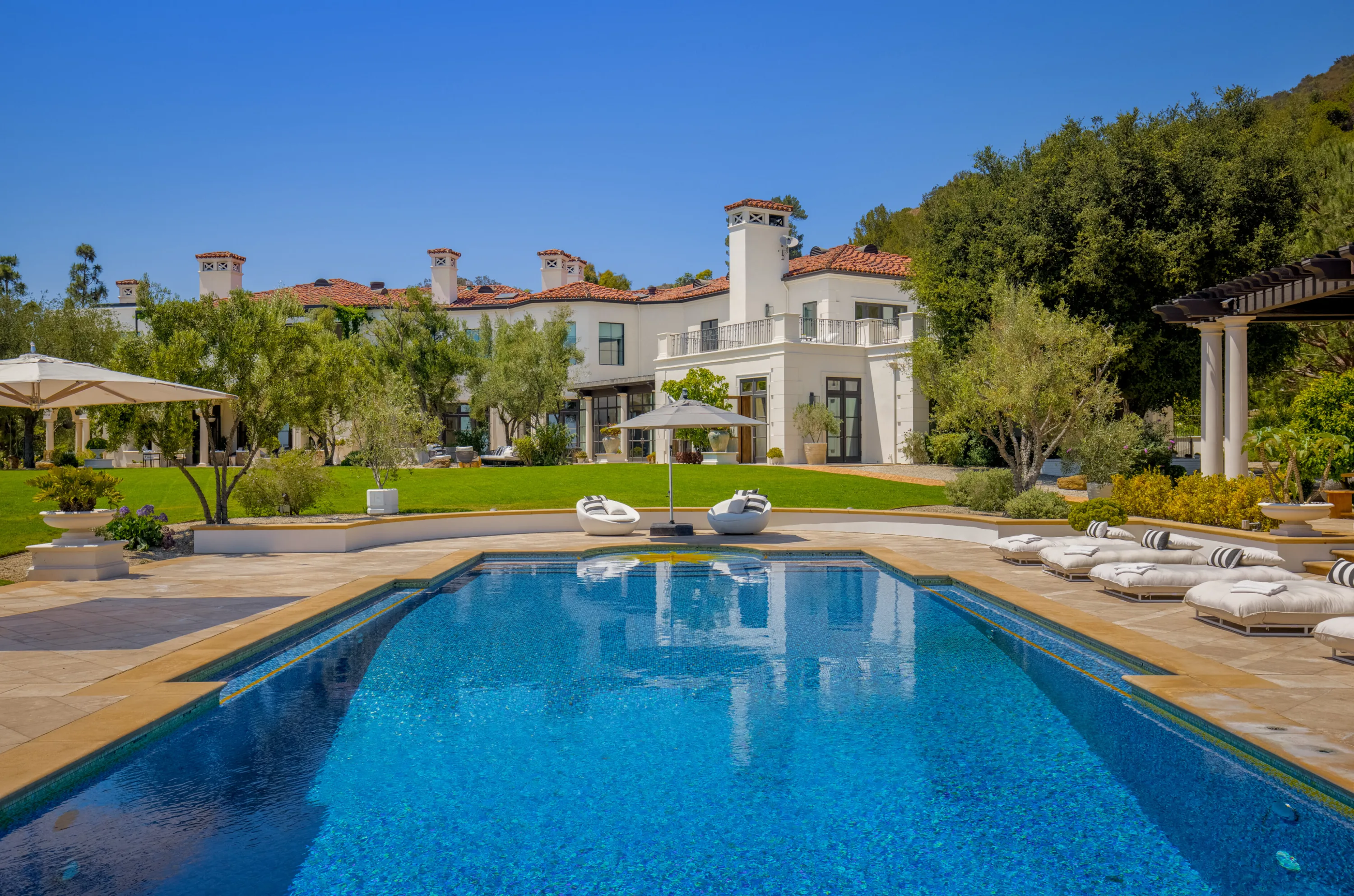 Wide pool with floating chaises and parasols, tiled roofs and chimneys of the estate rising beyond the lawn