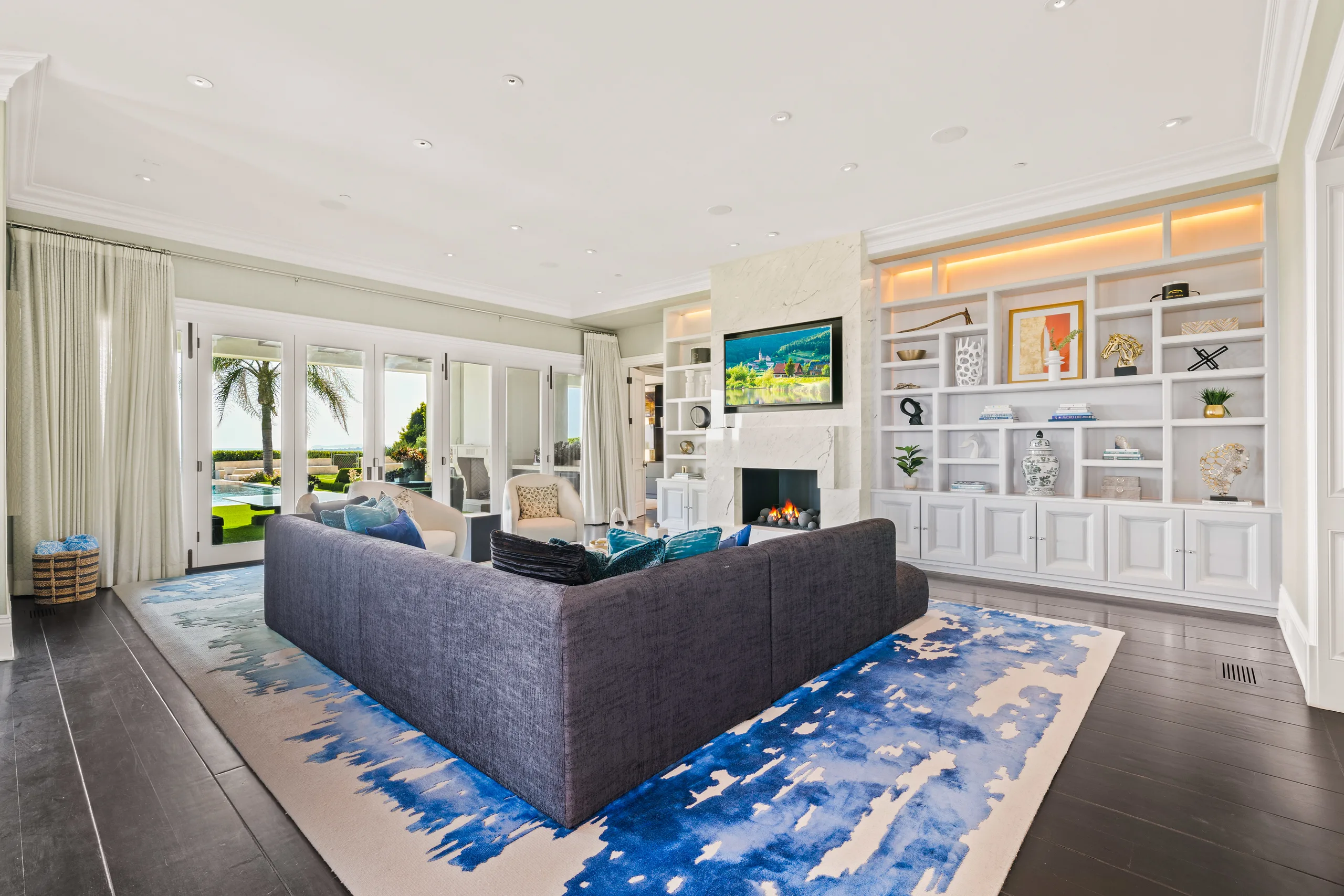 Same family room facing the fireplace, mounted screen and lit built-in shelving, pool glimpsed outside