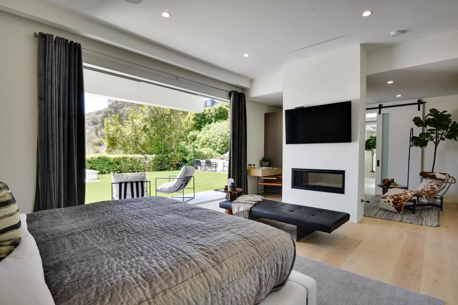 Primary suite with linear fireplace, wall-mounted TV and doors sliding open to the lawn terrace