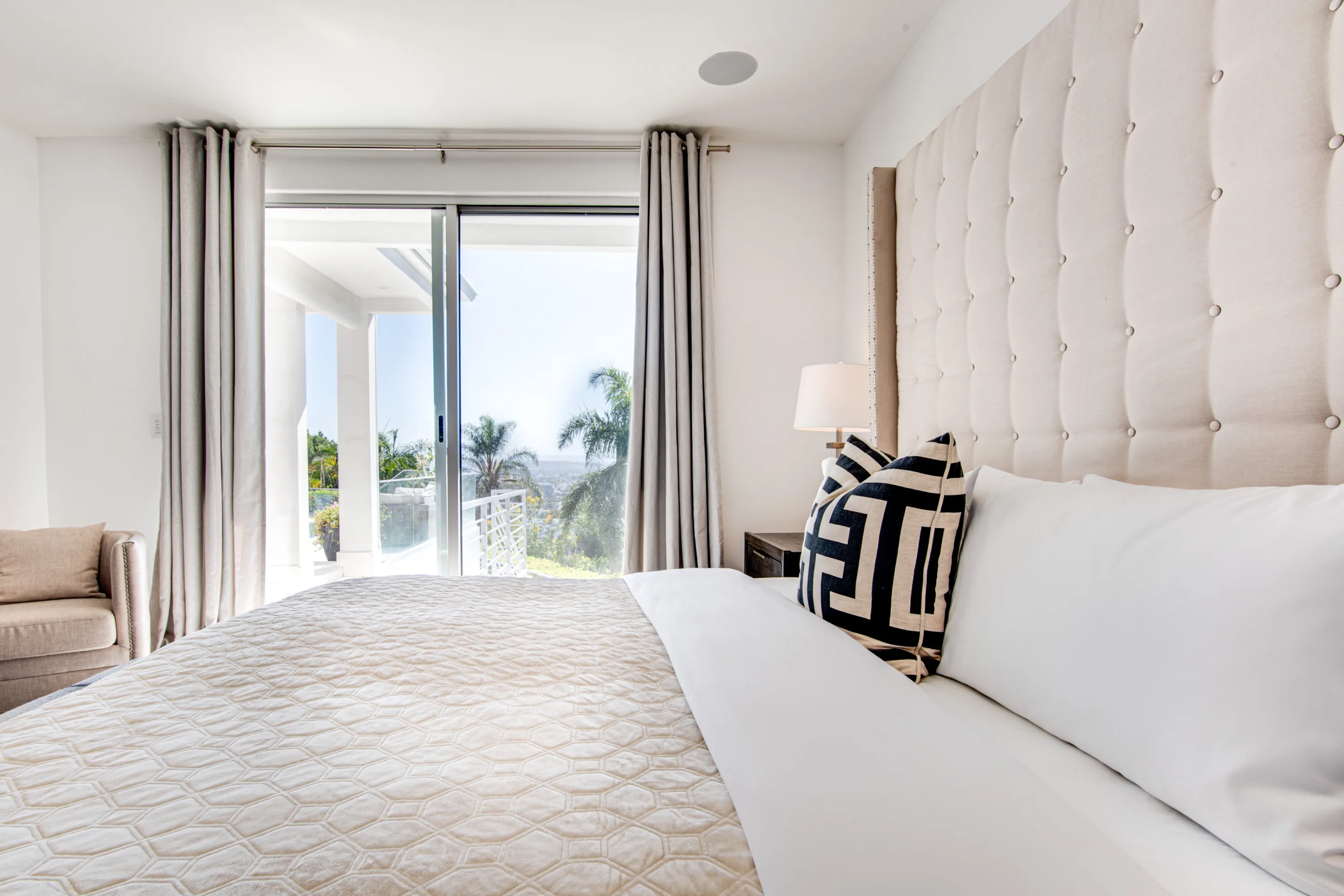 Bedside outlook through sliding glass to a palm-lined terrace and hazy city horizon