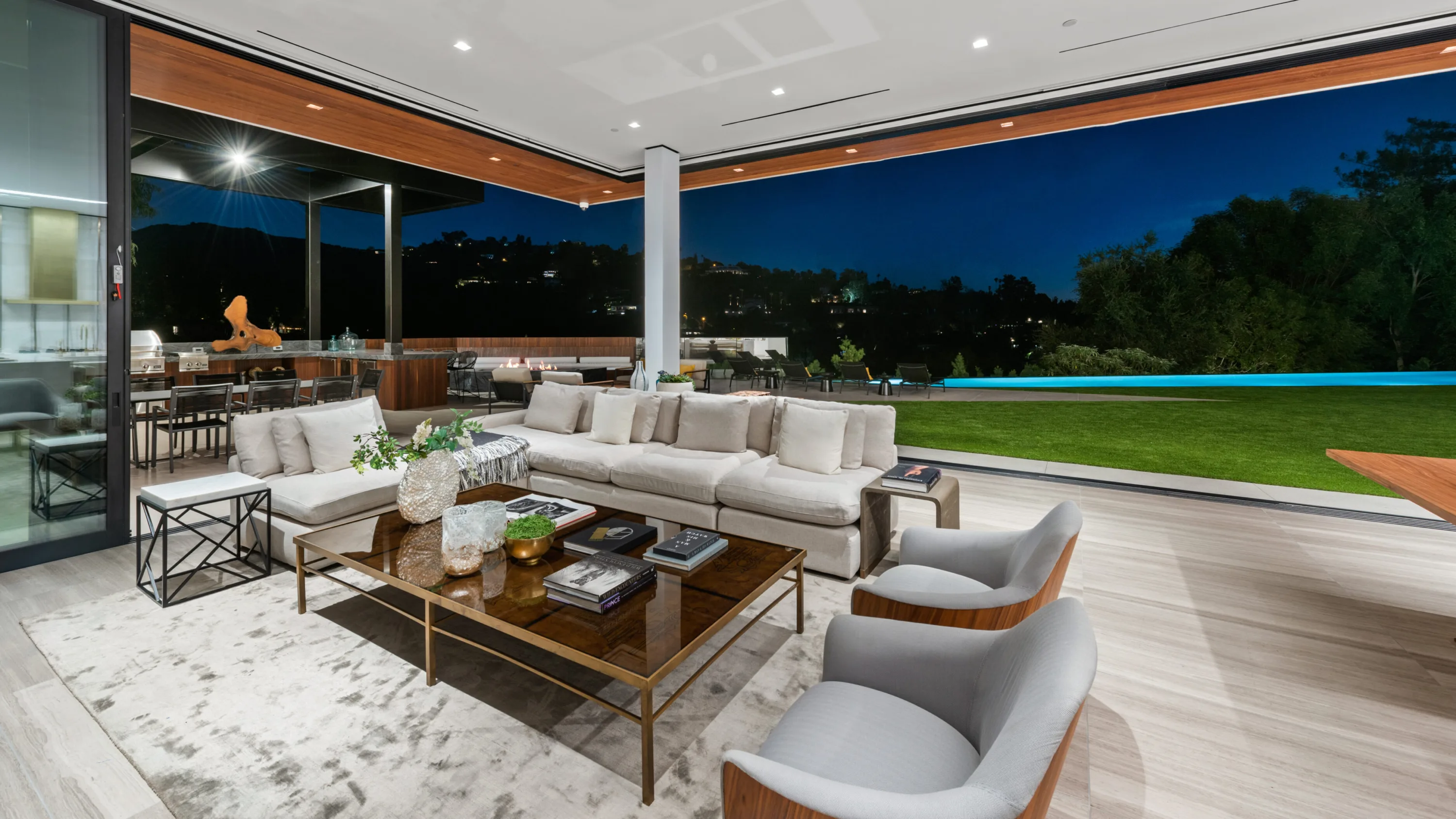 Evening in the pocketing living room, sectional facing the lit lawn, pool edge and hillside lights