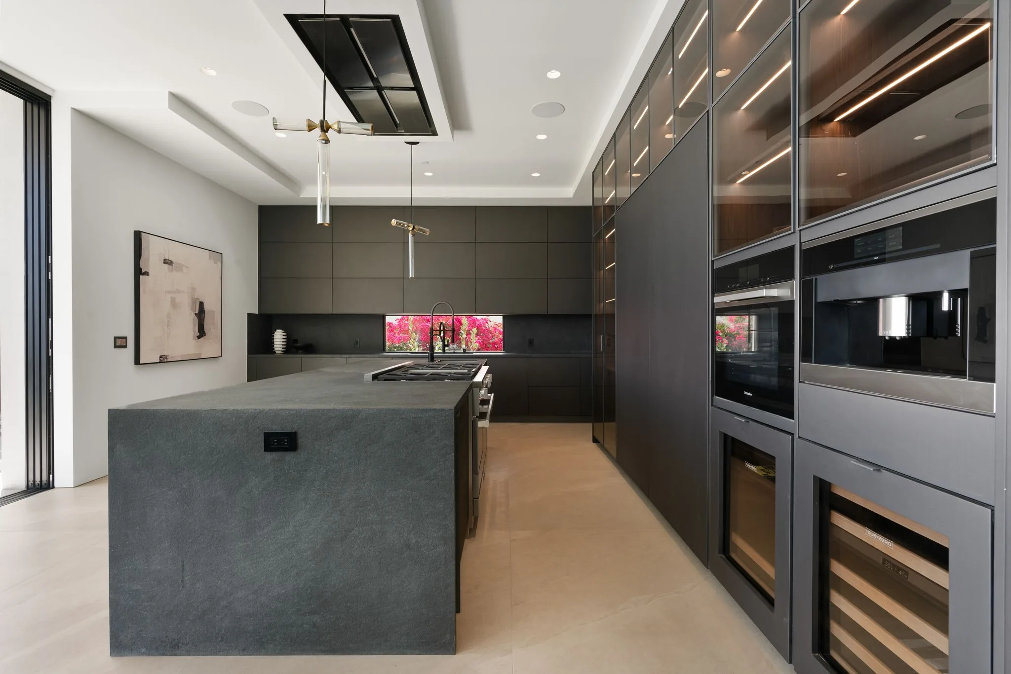 Chef's kitchen with a monolithic stone island, integrated ovens and a bougainvillea-framed window splashback