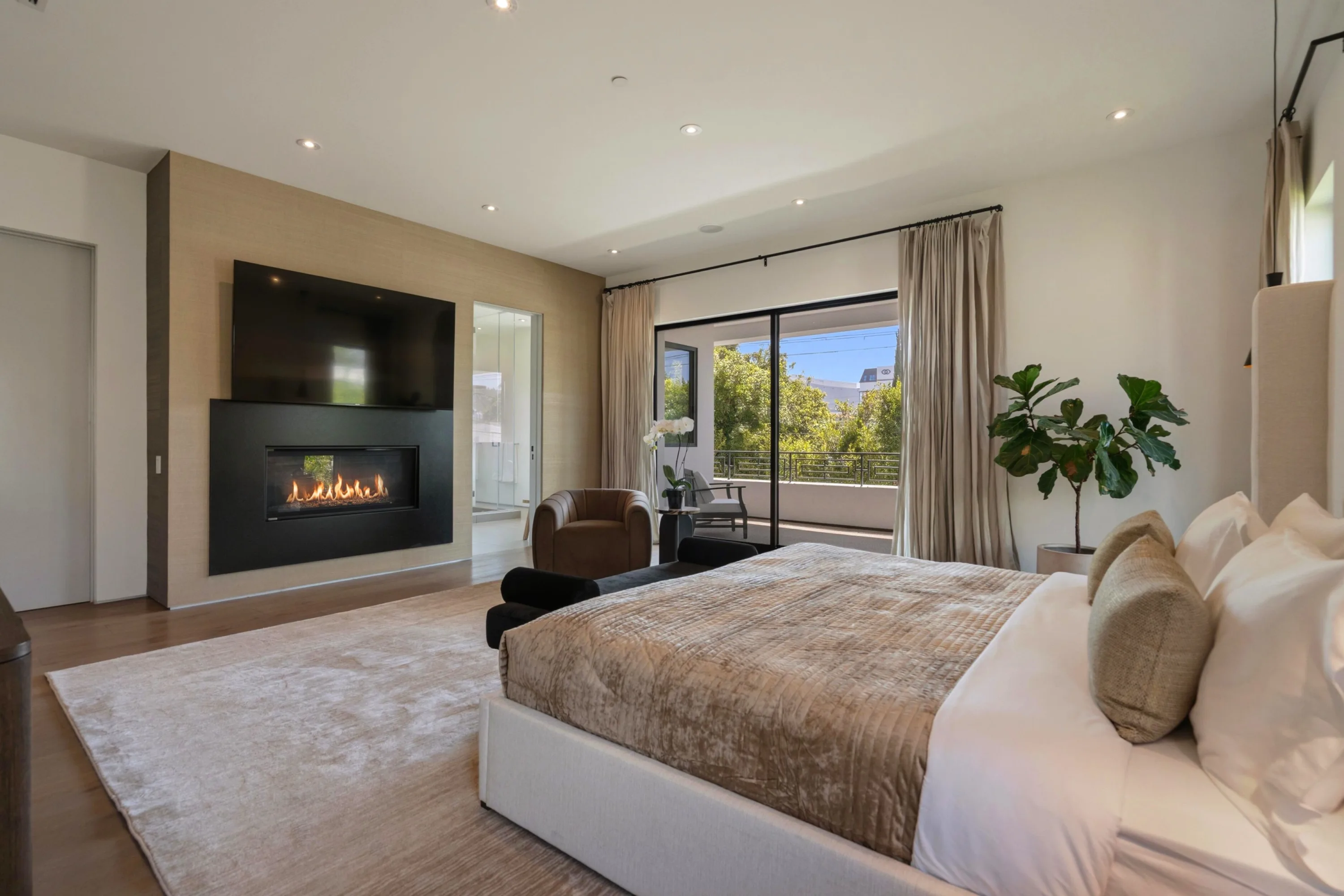 Second view of the primary bedroom, fireplace alight and curtains open to a balcony and city treeline
