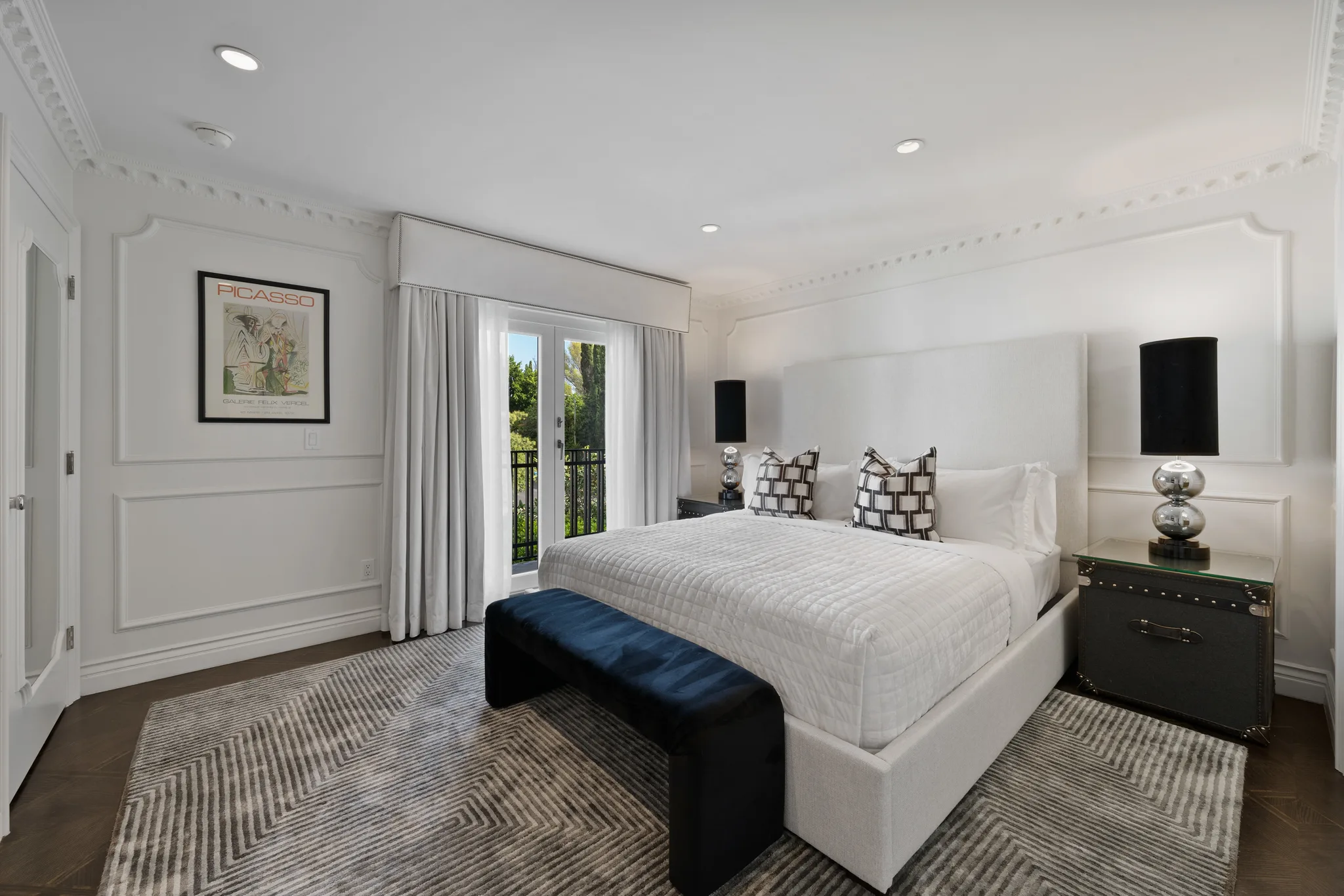 Guest bedroom with upholstered king bed, navy velvet bench and French doors to a balcony; framed Picasso exhibition poster on the panelled wall