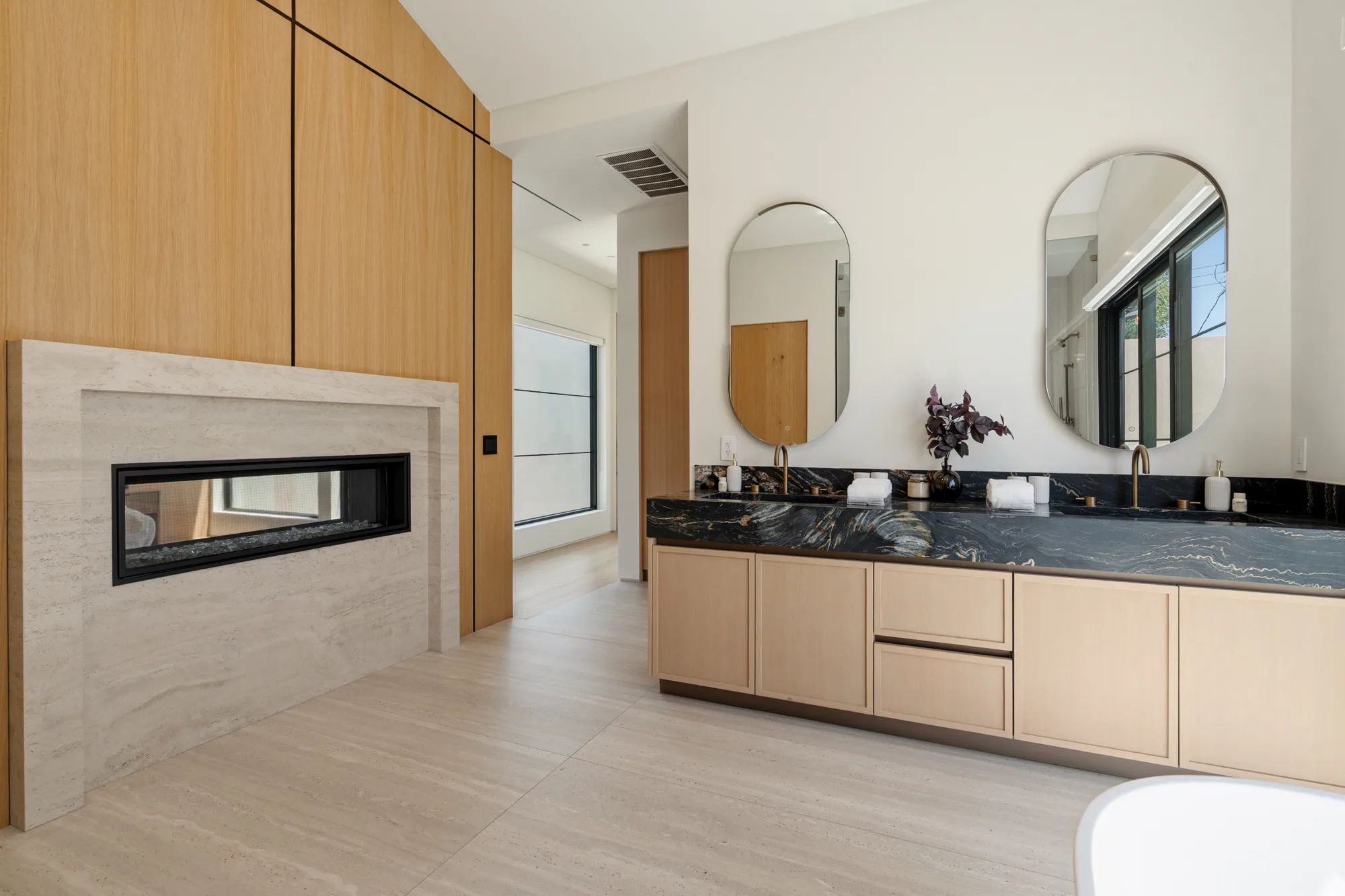 Primary bath with double vanity, dark veined stone counter, oval mirrors and a two-sided fireplace