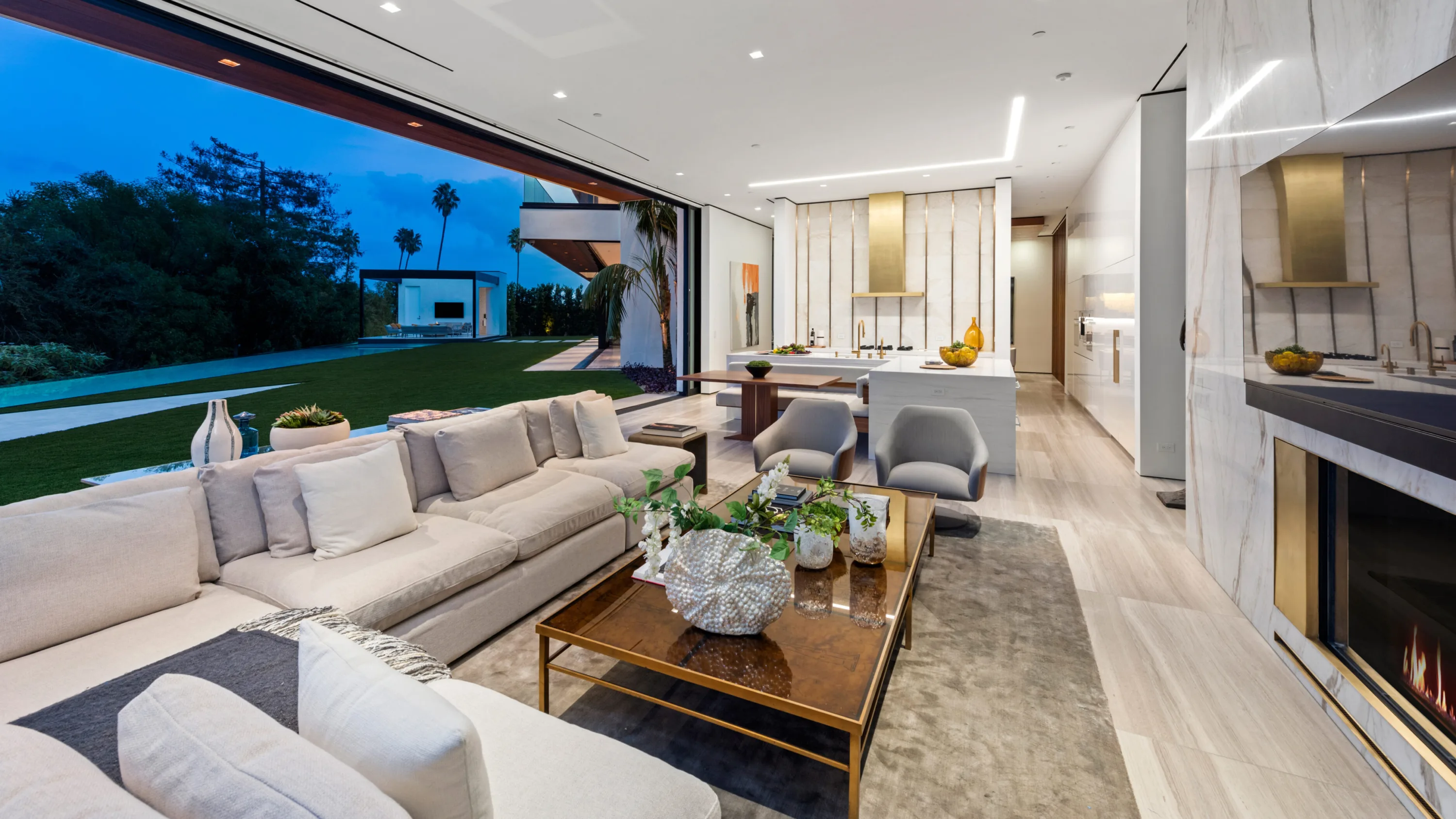 Sectional and fireplace with the kitchen beyond, glass wall retracted to lawn and poolside pavilion