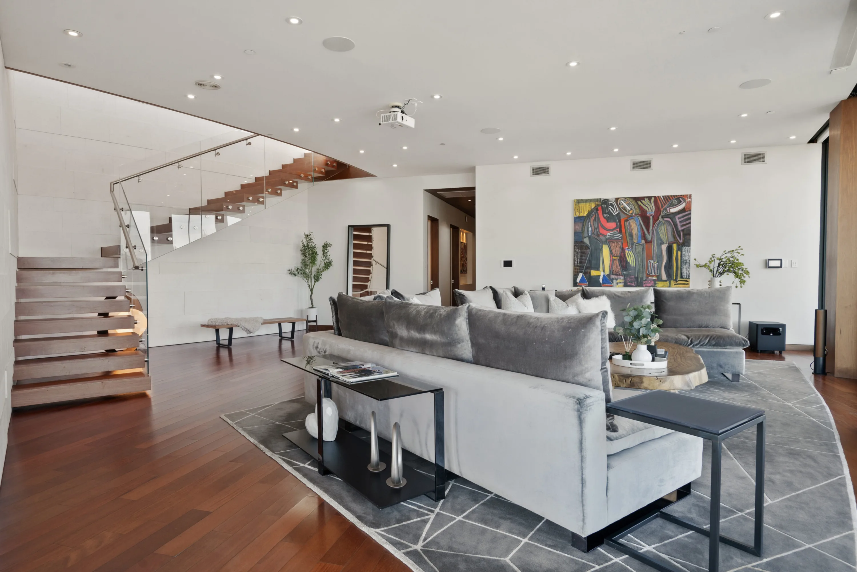 Great room with floating staircase behind glass balustrade, grey sofas and a large abstract canvas