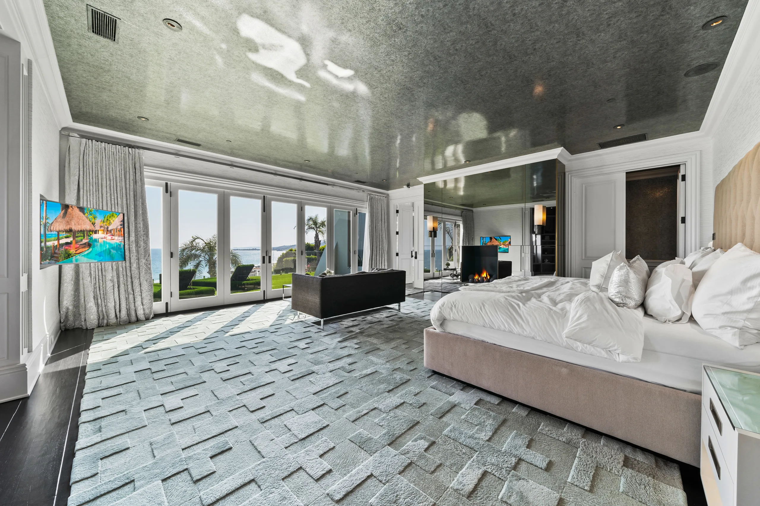 Primary suite from the bed side, glass doors framing lawn and sea, fire lit behind the seating area