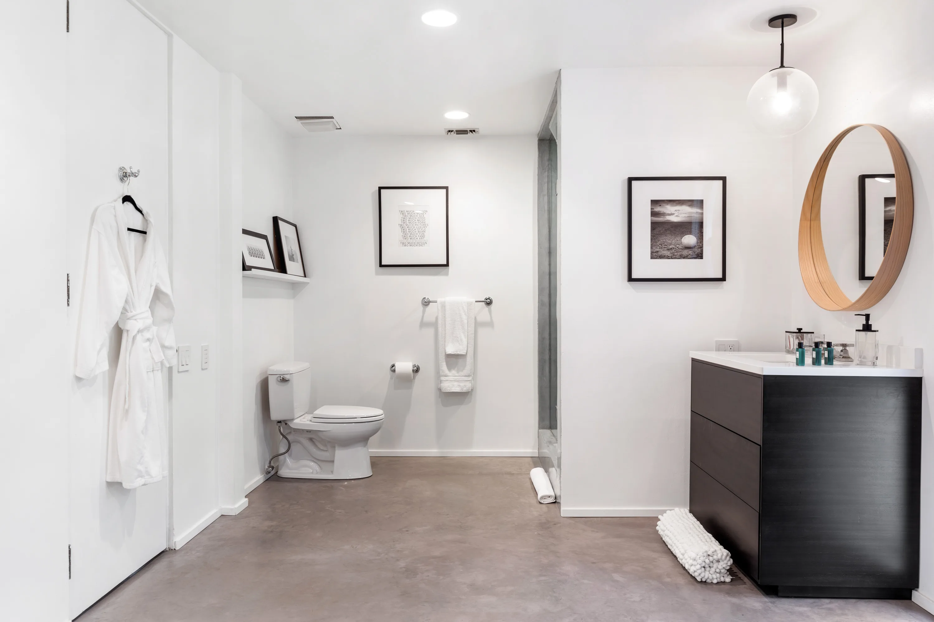 Bathroom in white with dark floating vanity, round mirror, robe on a hook and polished concrete floor