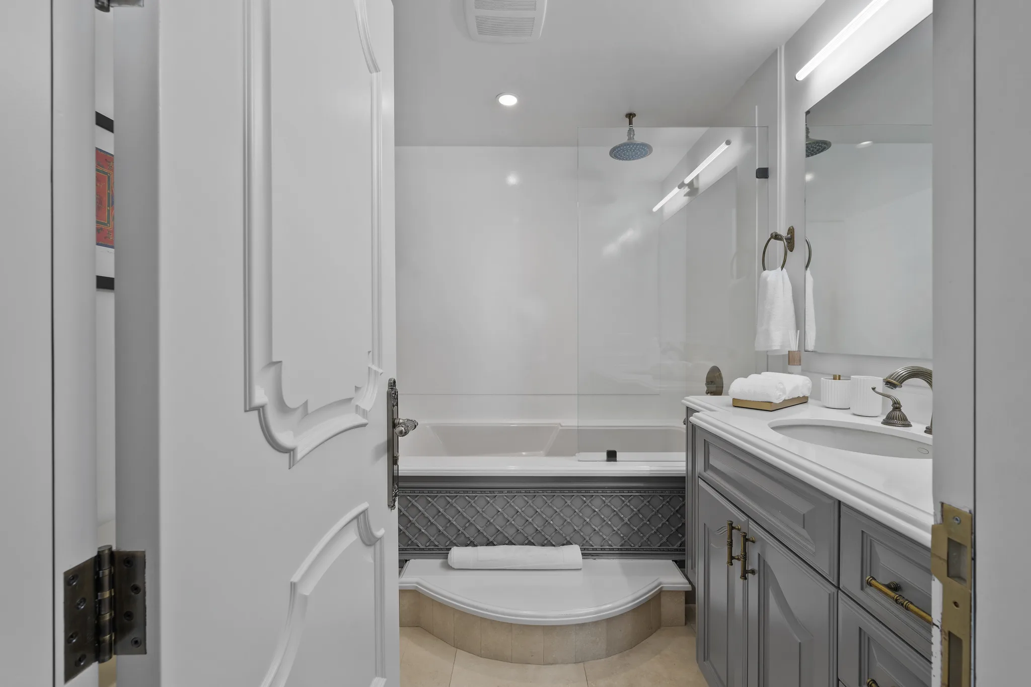 Ensuite bath seen through an open panelled door, with soaking tub under a rain shower and grey vanity with folded towels