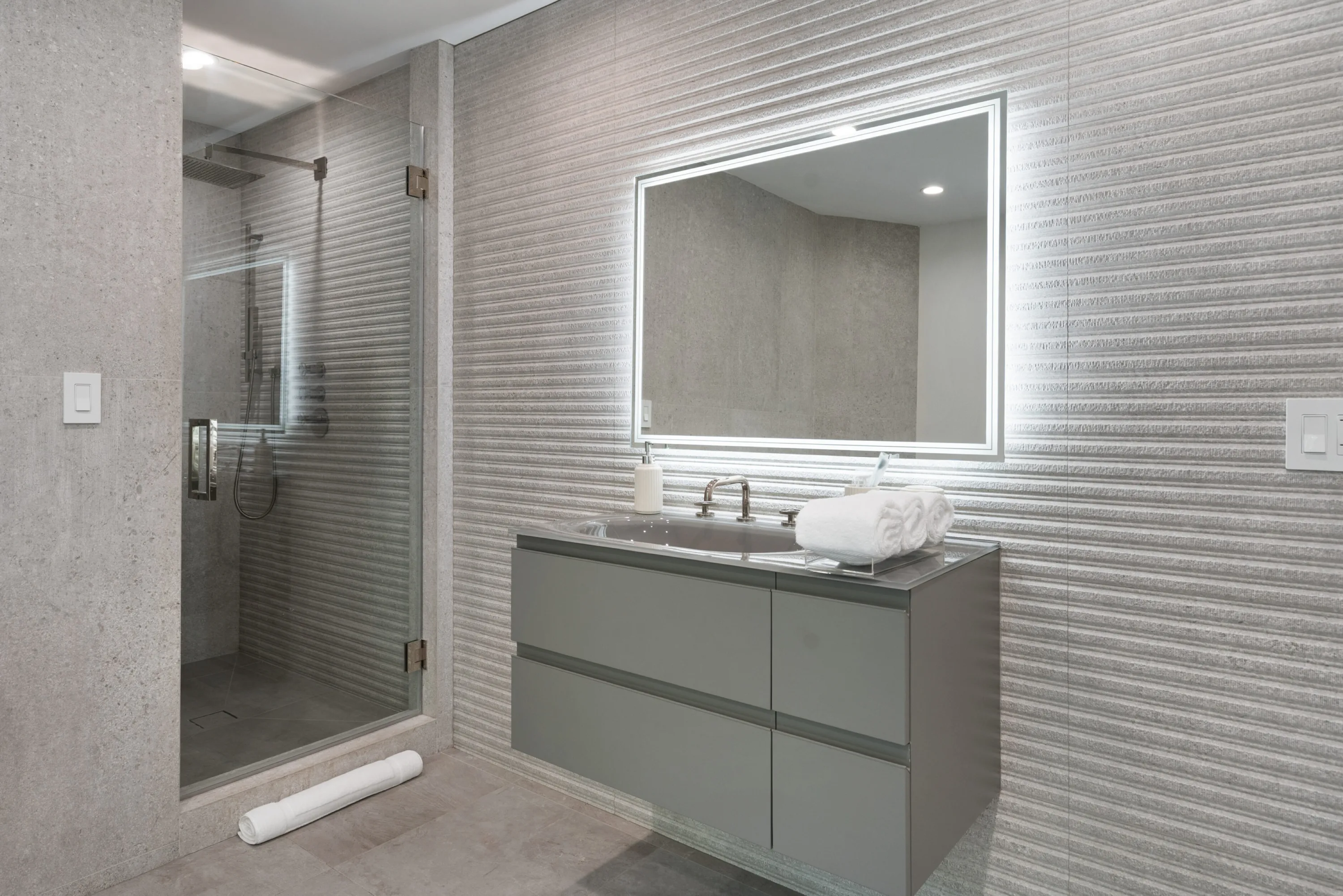 Bathroom clad in ribbed stone with walk-in shower and illuminated mirror over a grey vanity