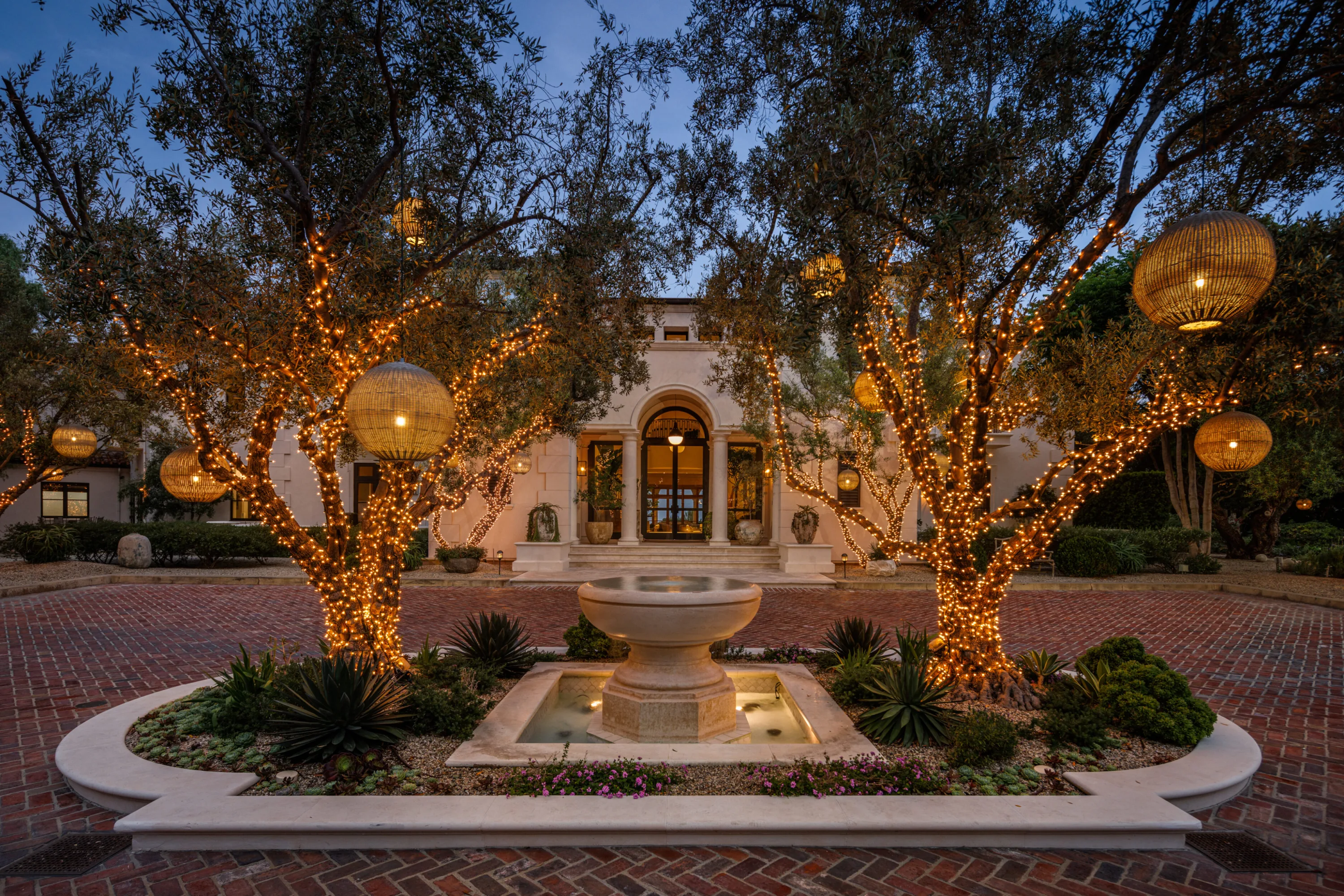 Stone fountain in the brick motor court, framed by fairy-lit olive trees and woven globe lanterns at twilight