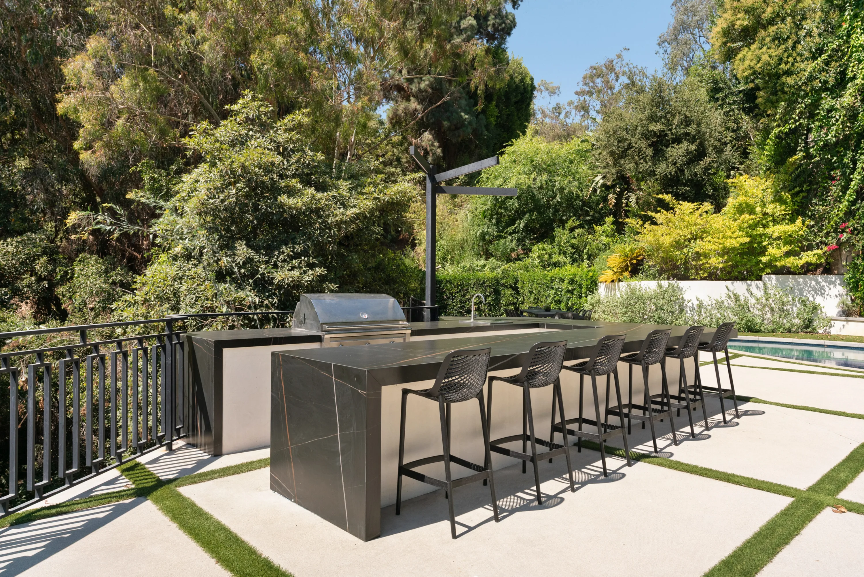 Outdoor kitchen with built-in grill, sink and six bar stools along a dark stone counter