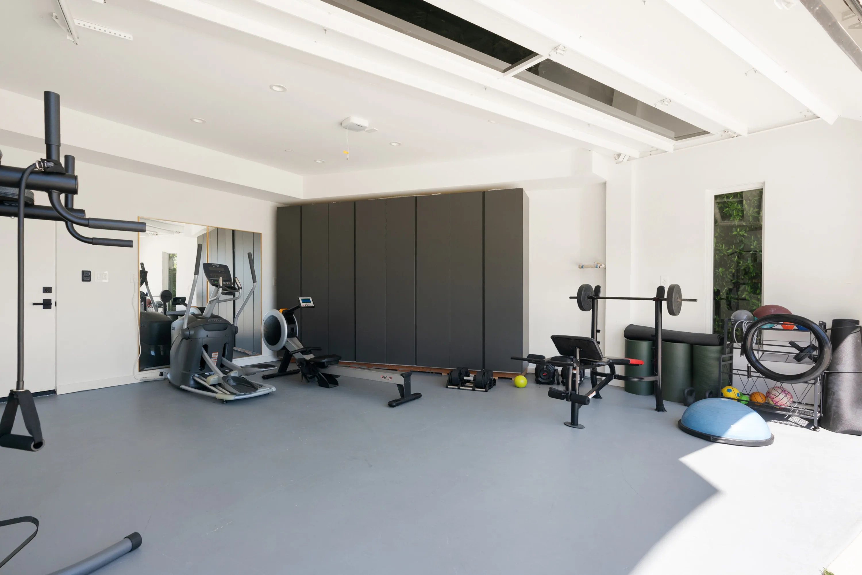 Home gym with elliptical, rower, weight bench, racked balls and a mirrored wall