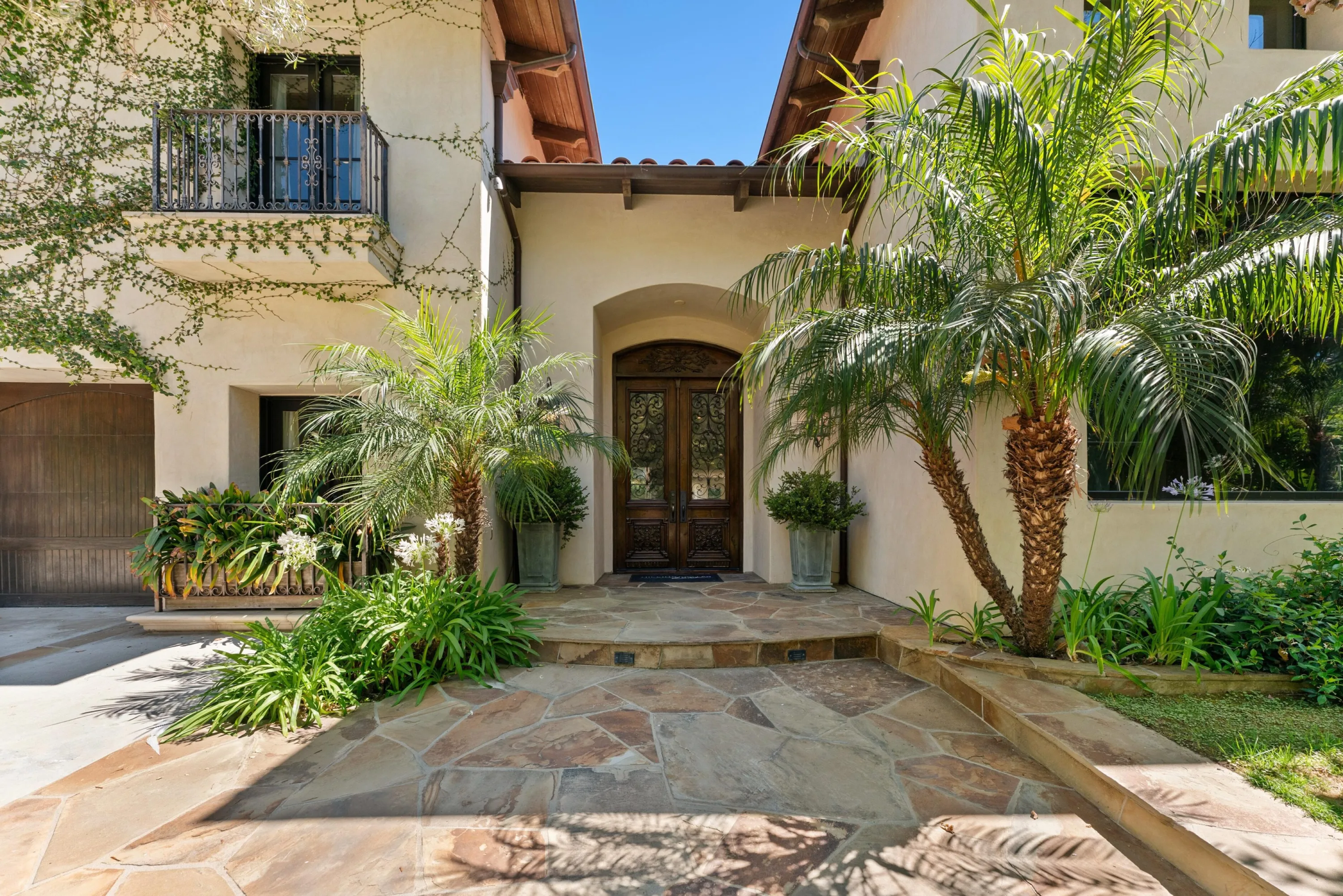 Flagstone entry path leading to carved double front doors framed by palms and potted greenery