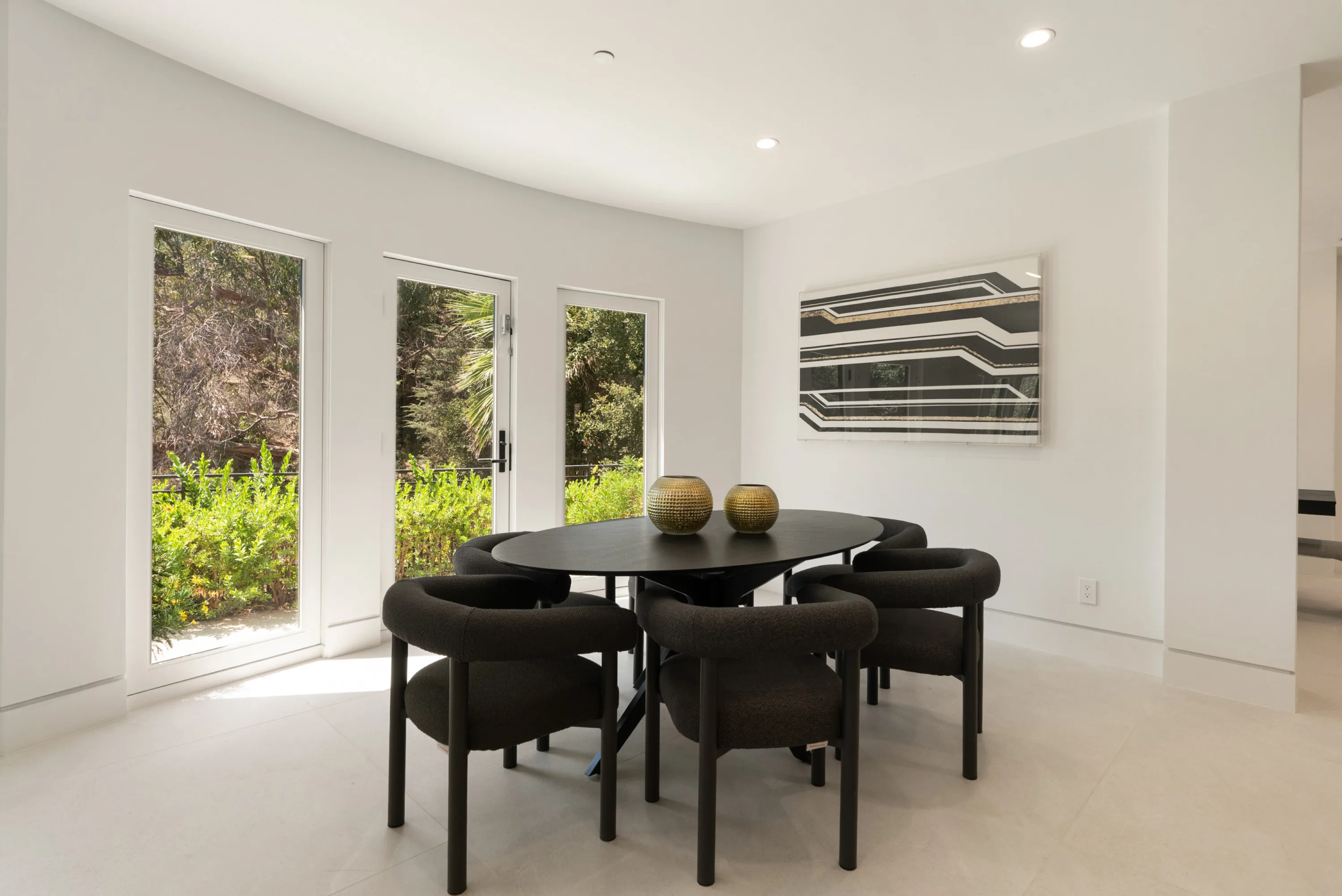 Breakfast nook with oval black table, six rounded chairs and hillside greenery through tall doors