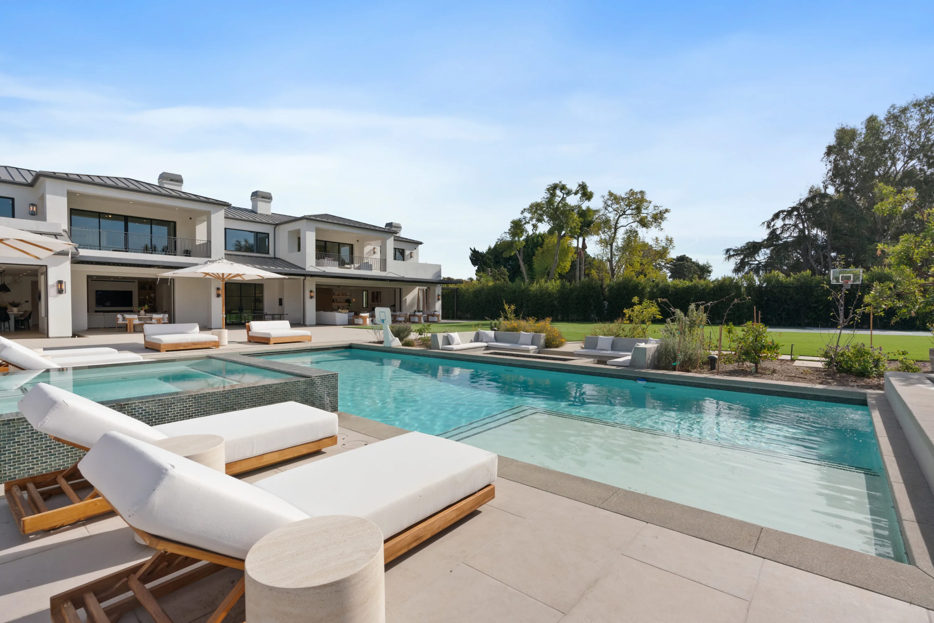 Poolside terrace with white teak daybeds, raised spa and shaded loggia, a basketball hoop on the lawn beyond