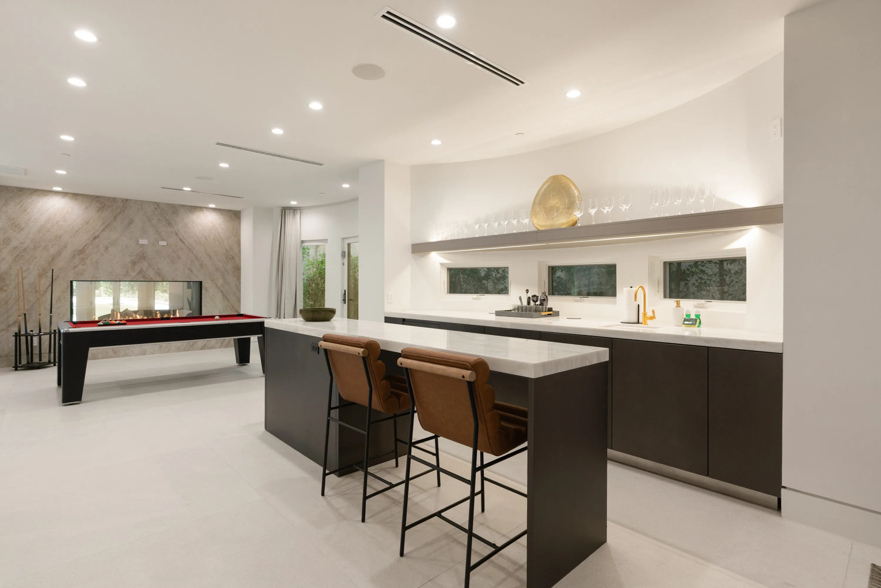 Entertaining level wet bar with stone counter and leather stools, pool table and fireplace beyond