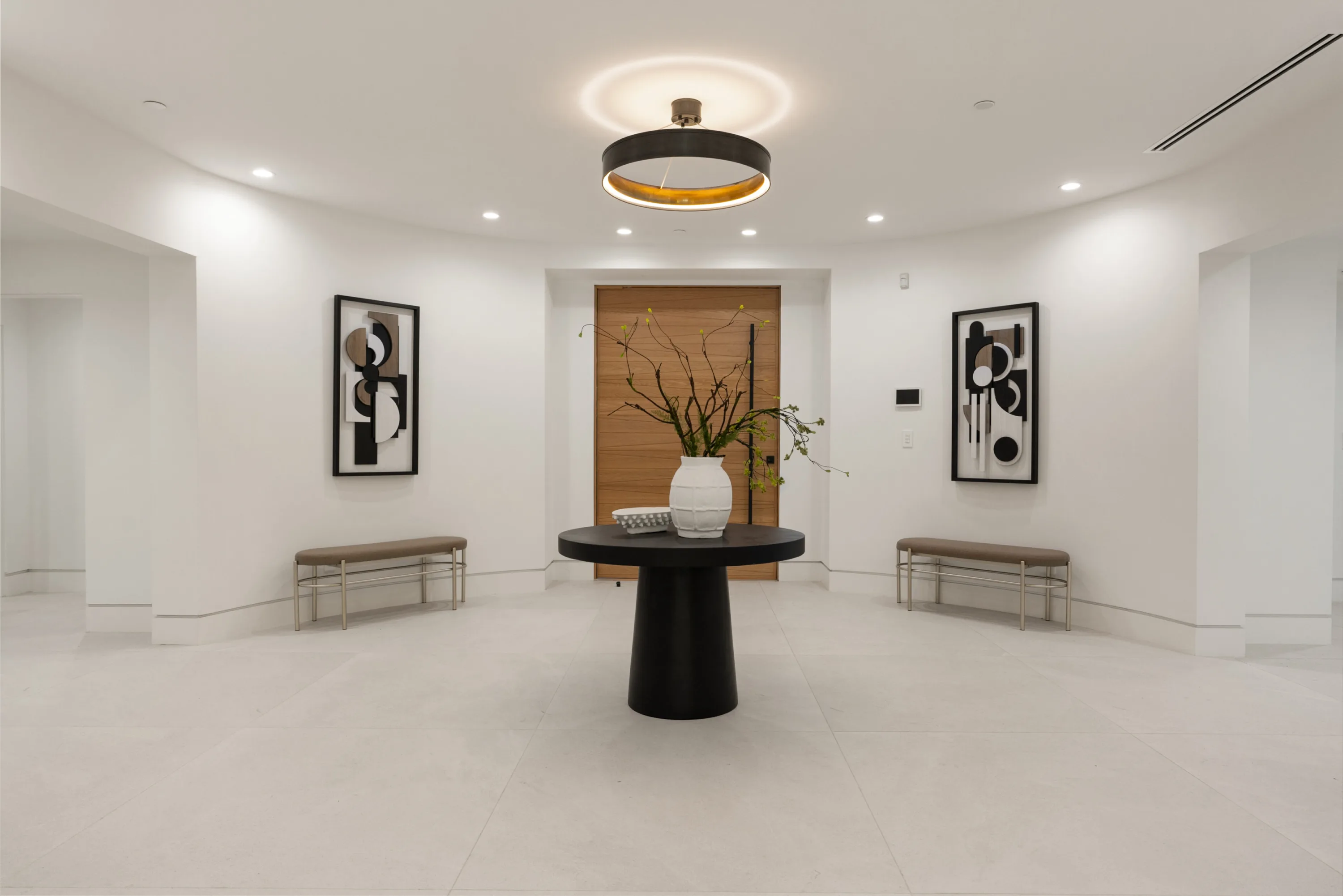 Rounded entry vestibule with black pedestal table, paired benches and graphic artwork flanking the oak door