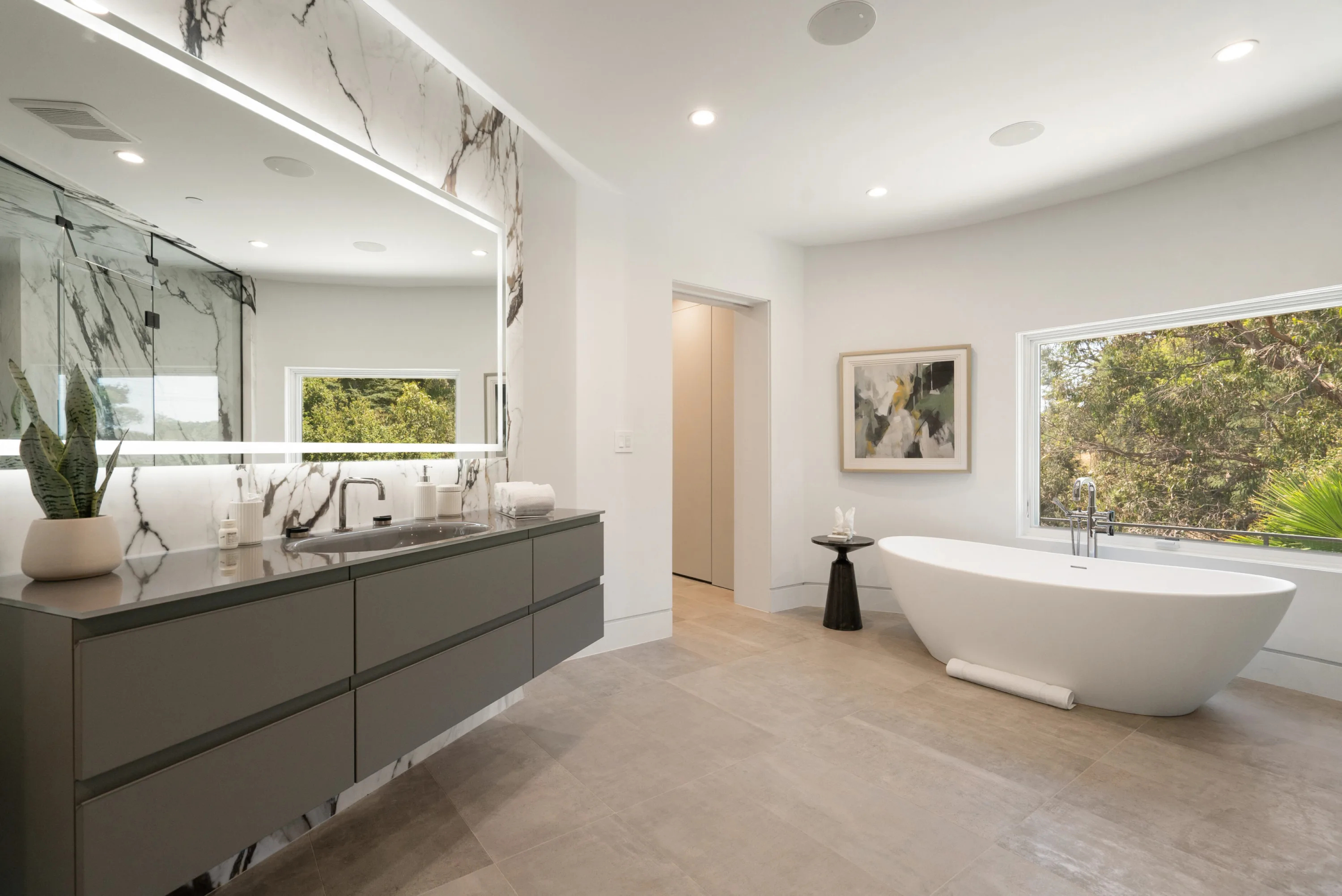 Primary bath with backlit mirror, floating double vanity and freestanding soaking tub beneath a window of green canopy