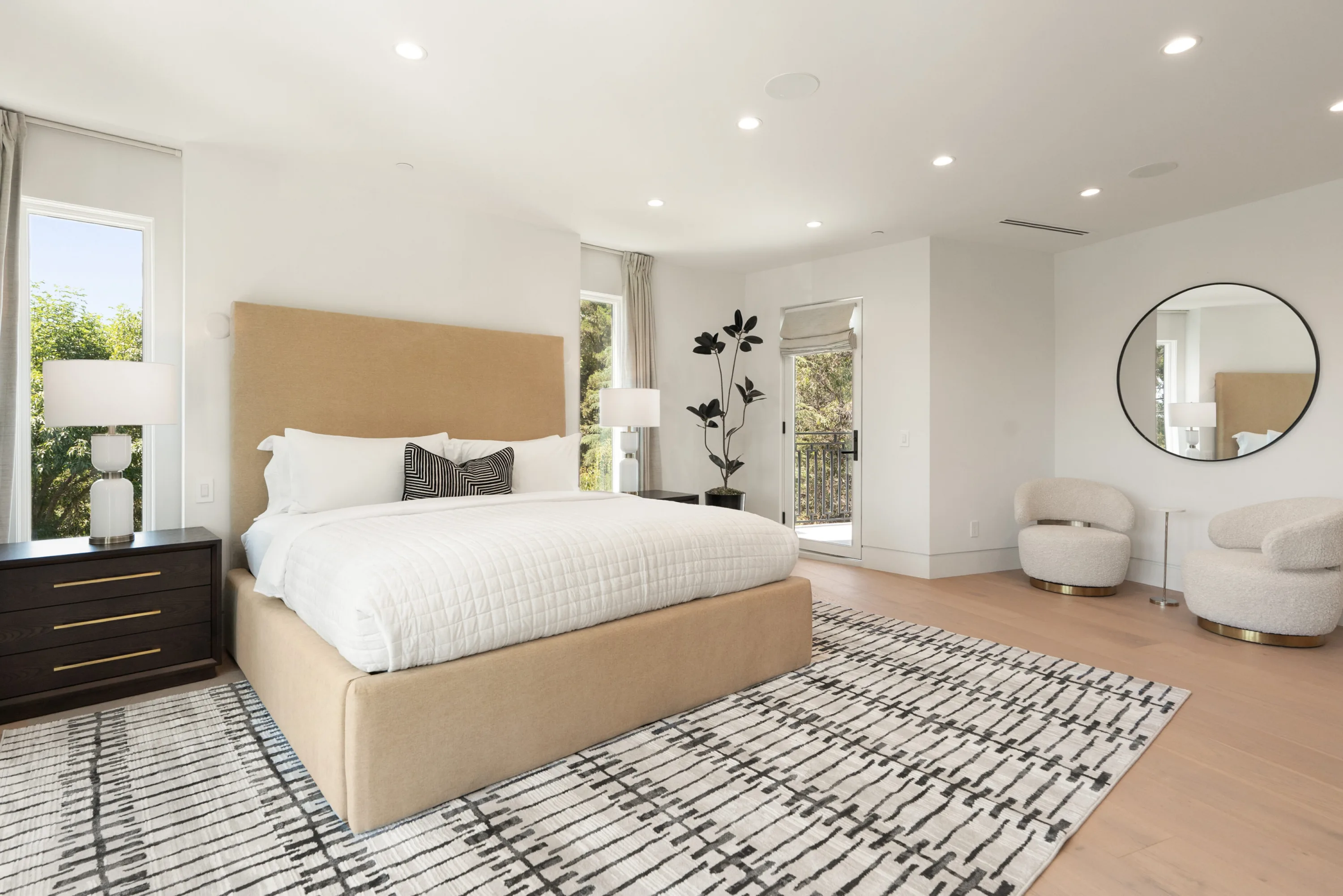Guest bedroom with tan upholstered bed, graphic rug, round mirror and doors to a small balcony