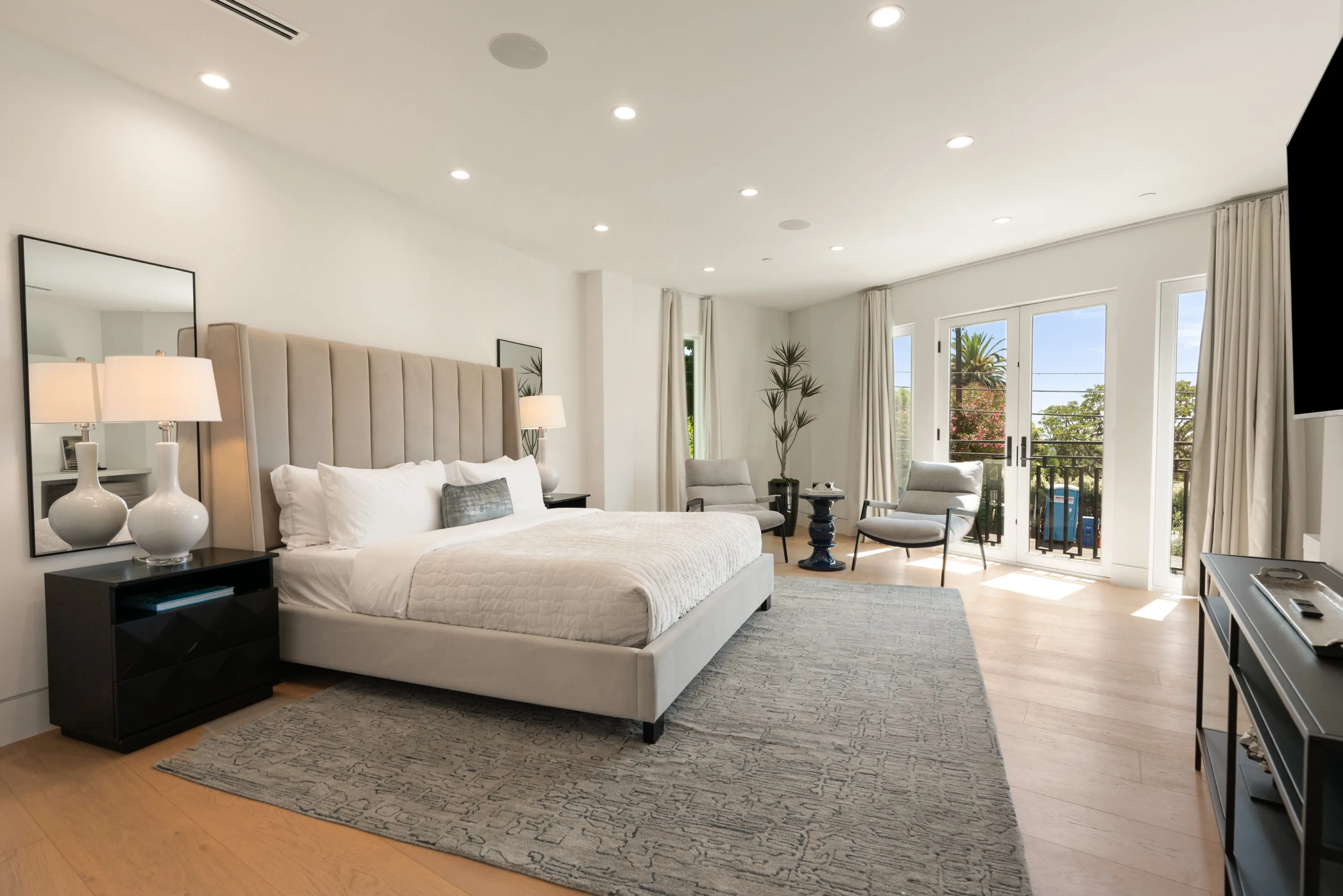 Airy guest suite with channel-tufted headboard, lounge chairs and French doors to a treetop balcony