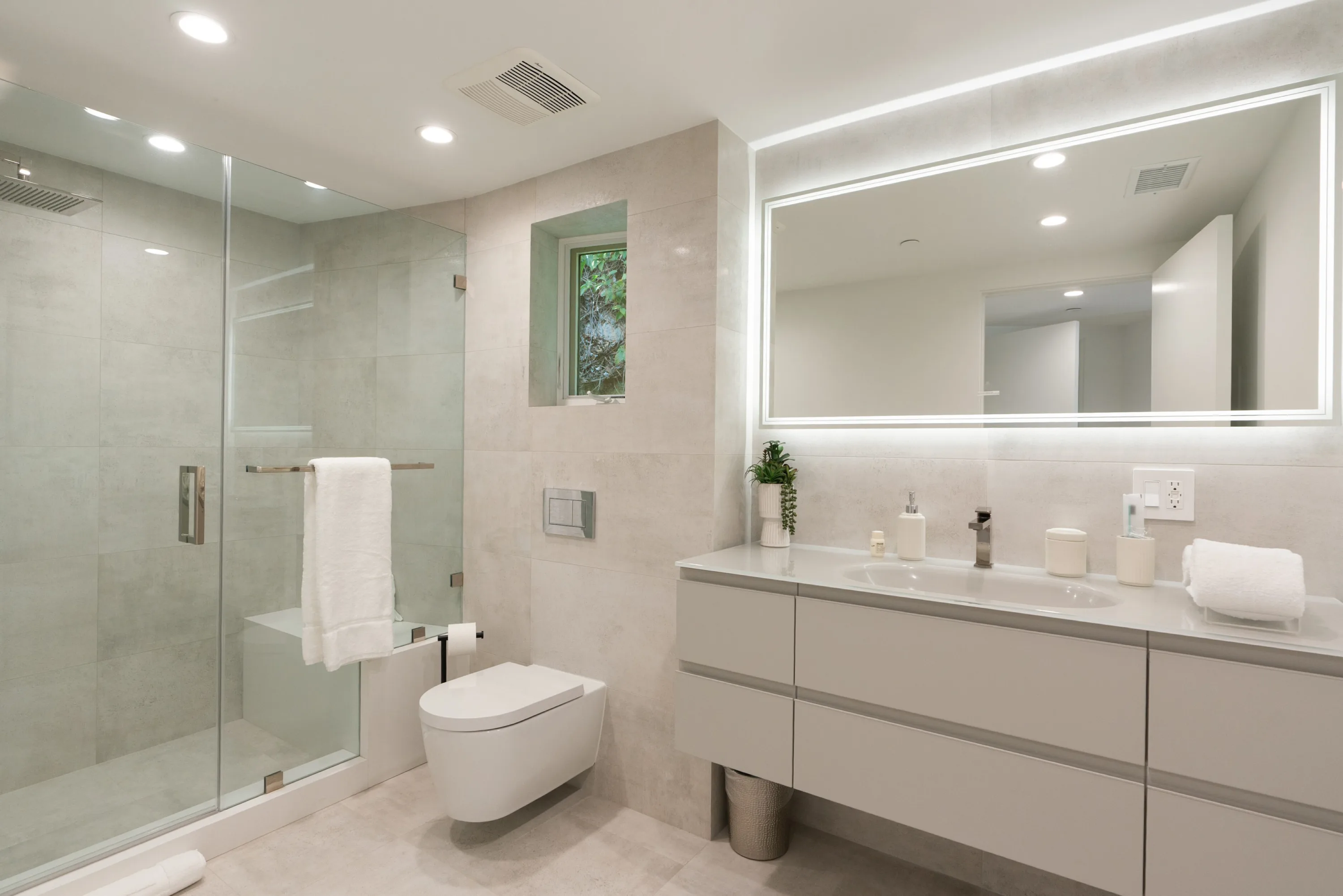 Ensuite bath in soft stone tile with walk-in glass shower, wall-hung basin and illuminated mirror