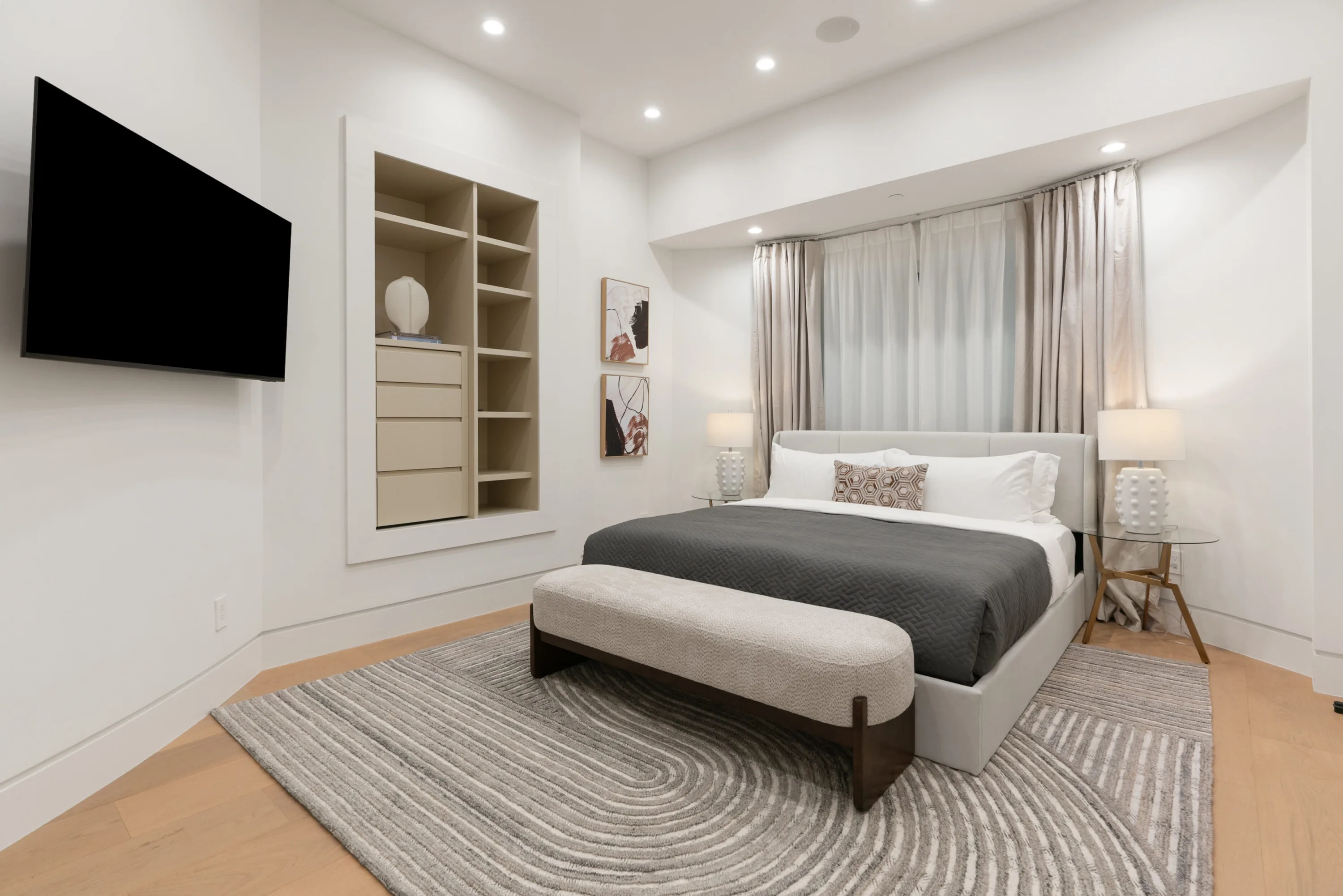 Guest bedroom with charcoal throw, upholstered bench and recessed shelving beneath a mounted TV
