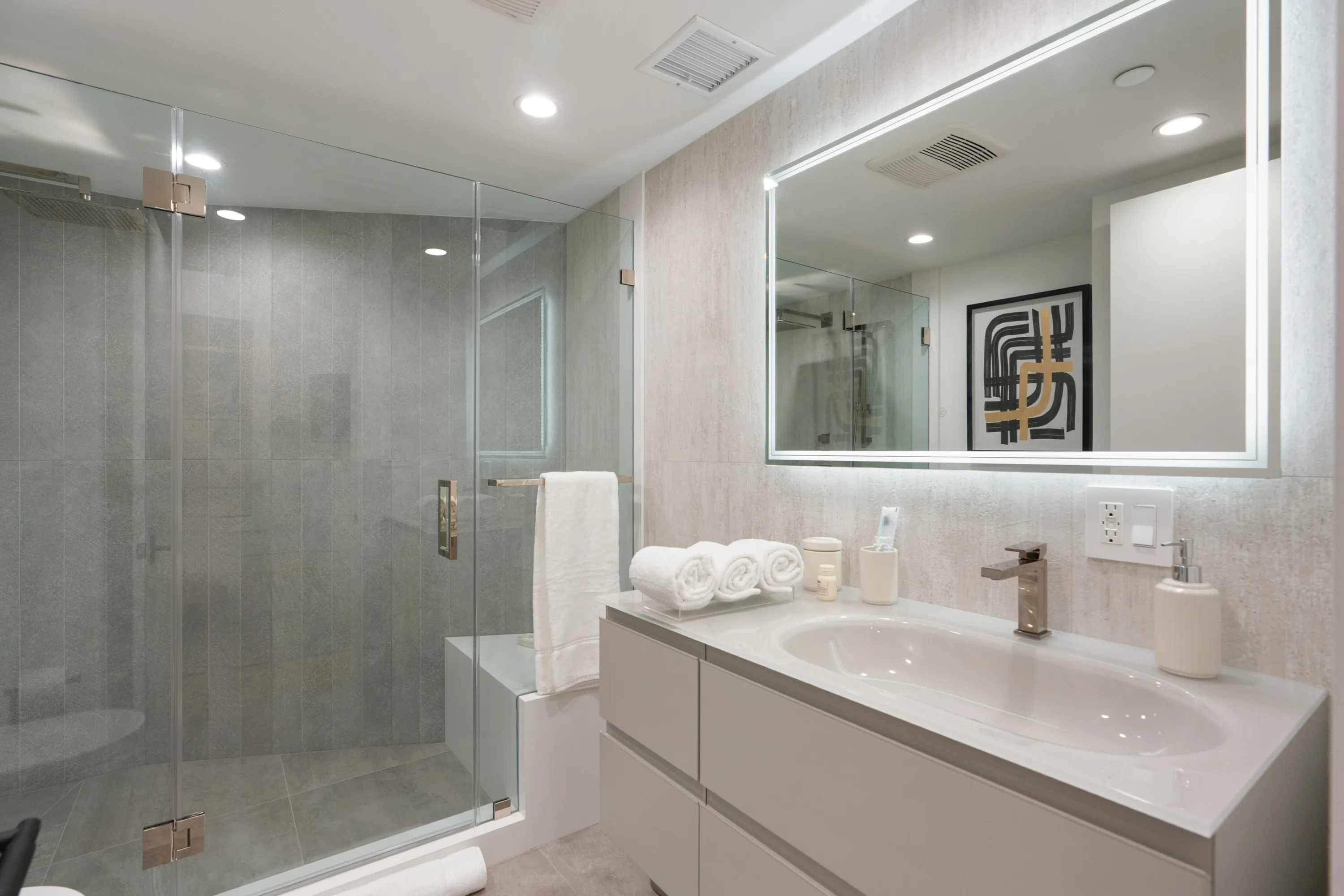 Tiled guest bath with oversized glass shower, rolled towels and a graphic print reflected in the lit mirror