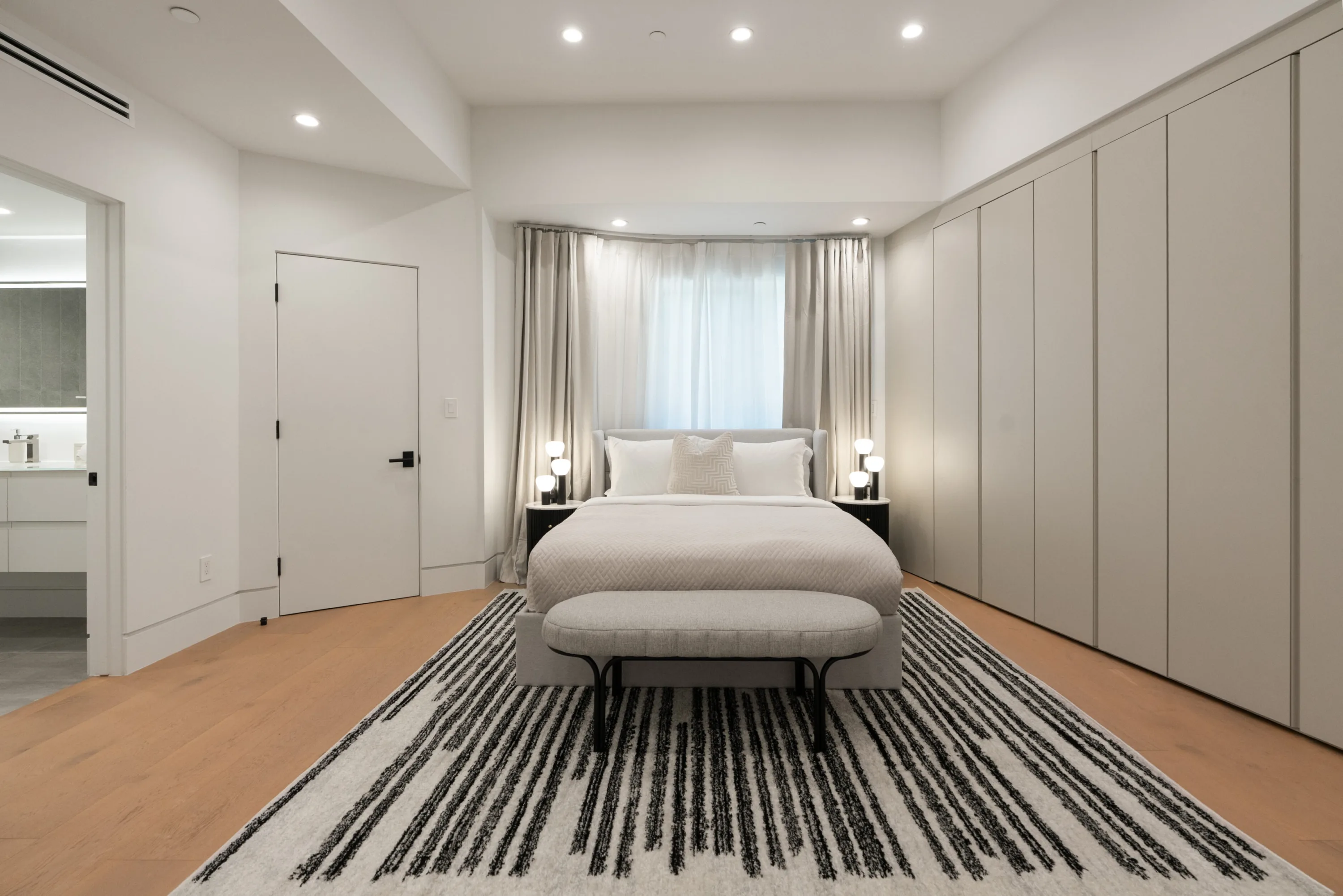Symmetrical bedroom with curtained bay window, bed bench and full wall of concealed closets