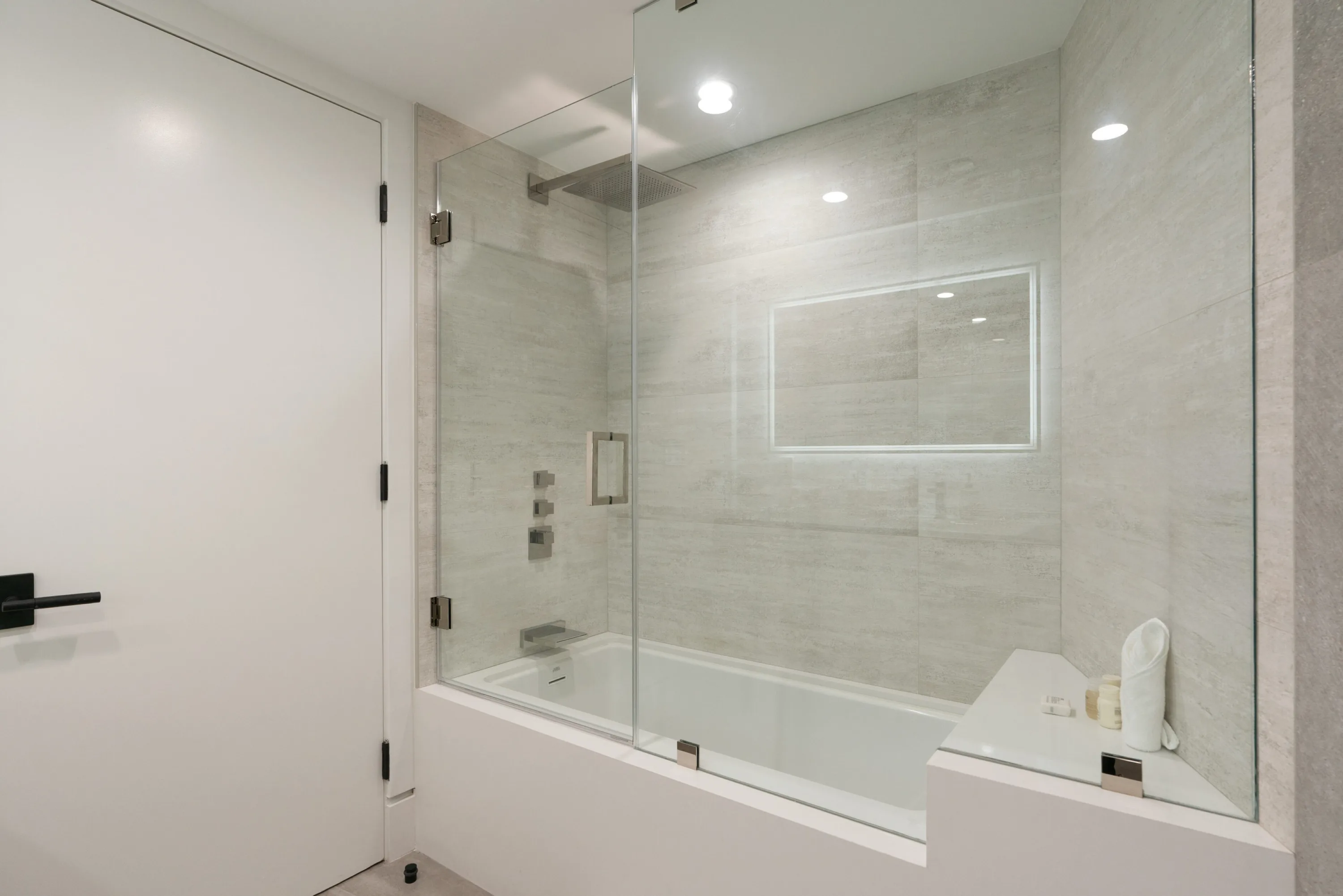 Soaking tub under a rain shower behind frameless glass, framed by pale travertine-look tile