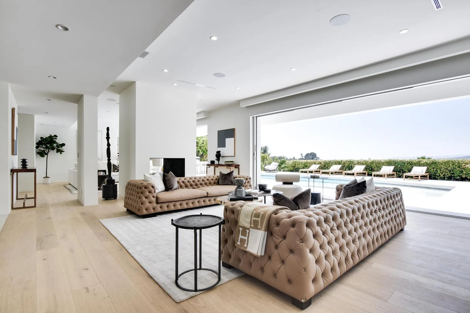 Twin tufted sofas on pale oak floors facing a wall of glass and the sun-lounger-lined pool deck