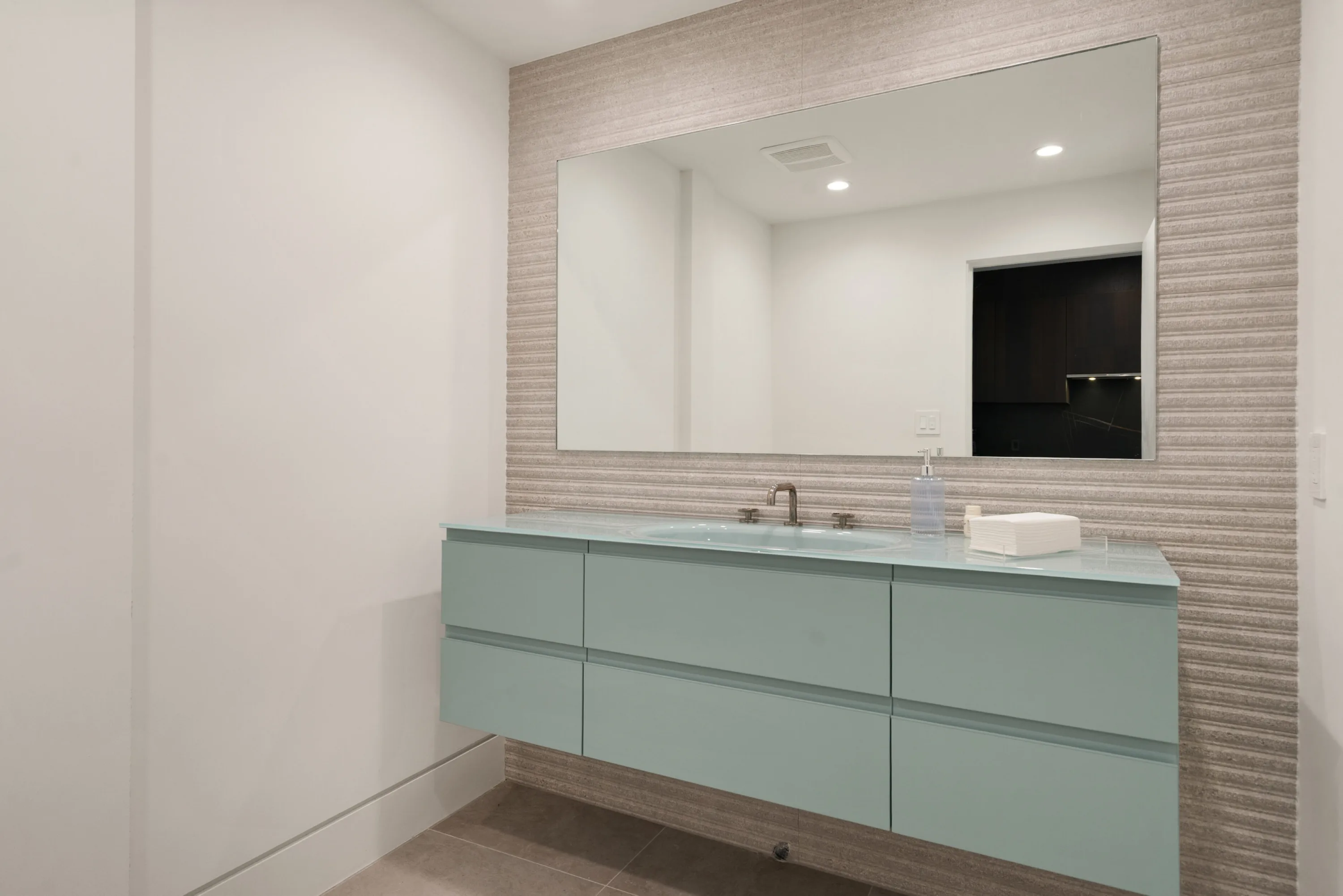 Powder room with seafoam floating vanity, glass countertop and wide mirror on ribbed stone