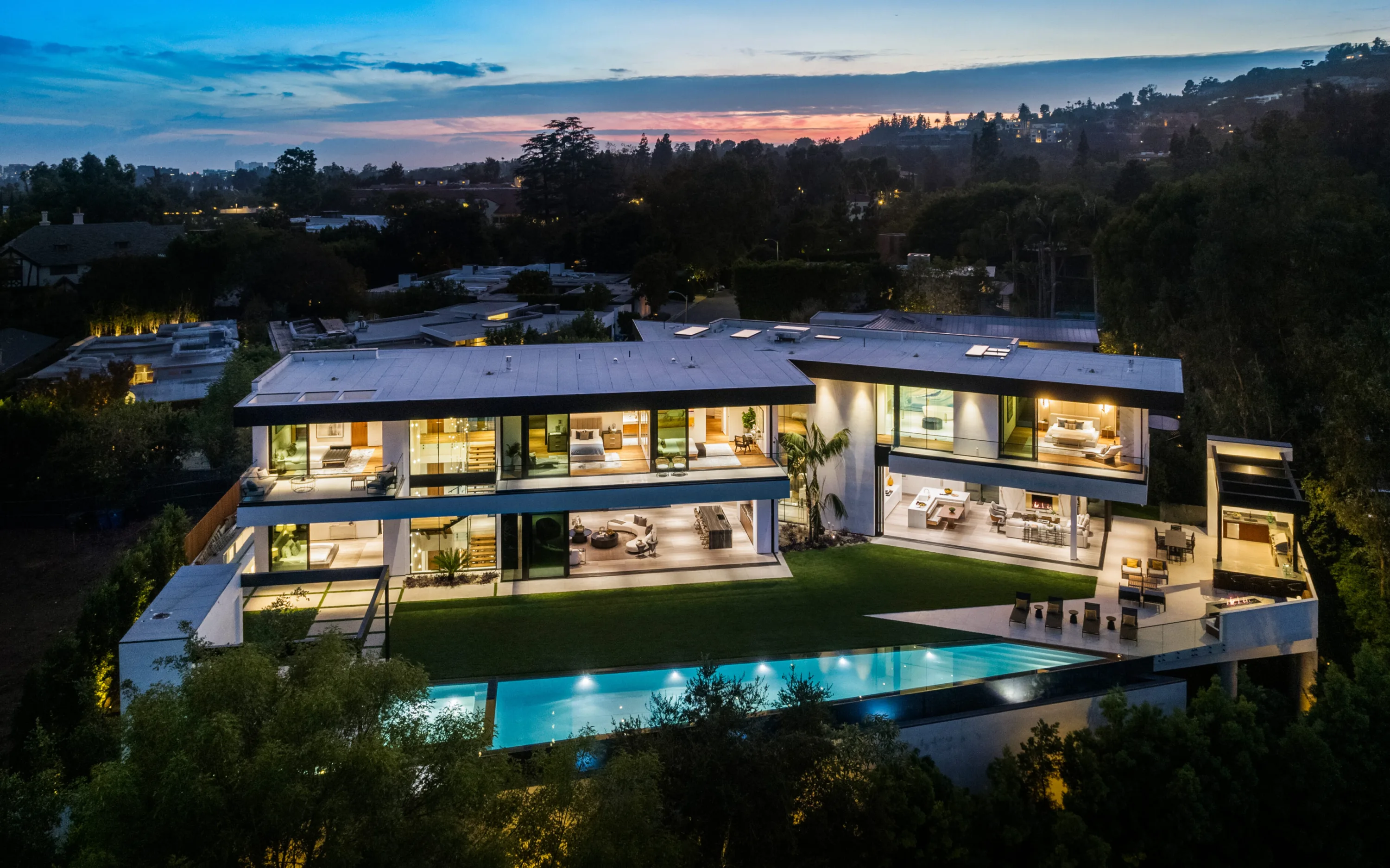 Aerial view at dusk of the glass-fronted Beverly Hills estate, lit rooms open to lawn and long infinity pool