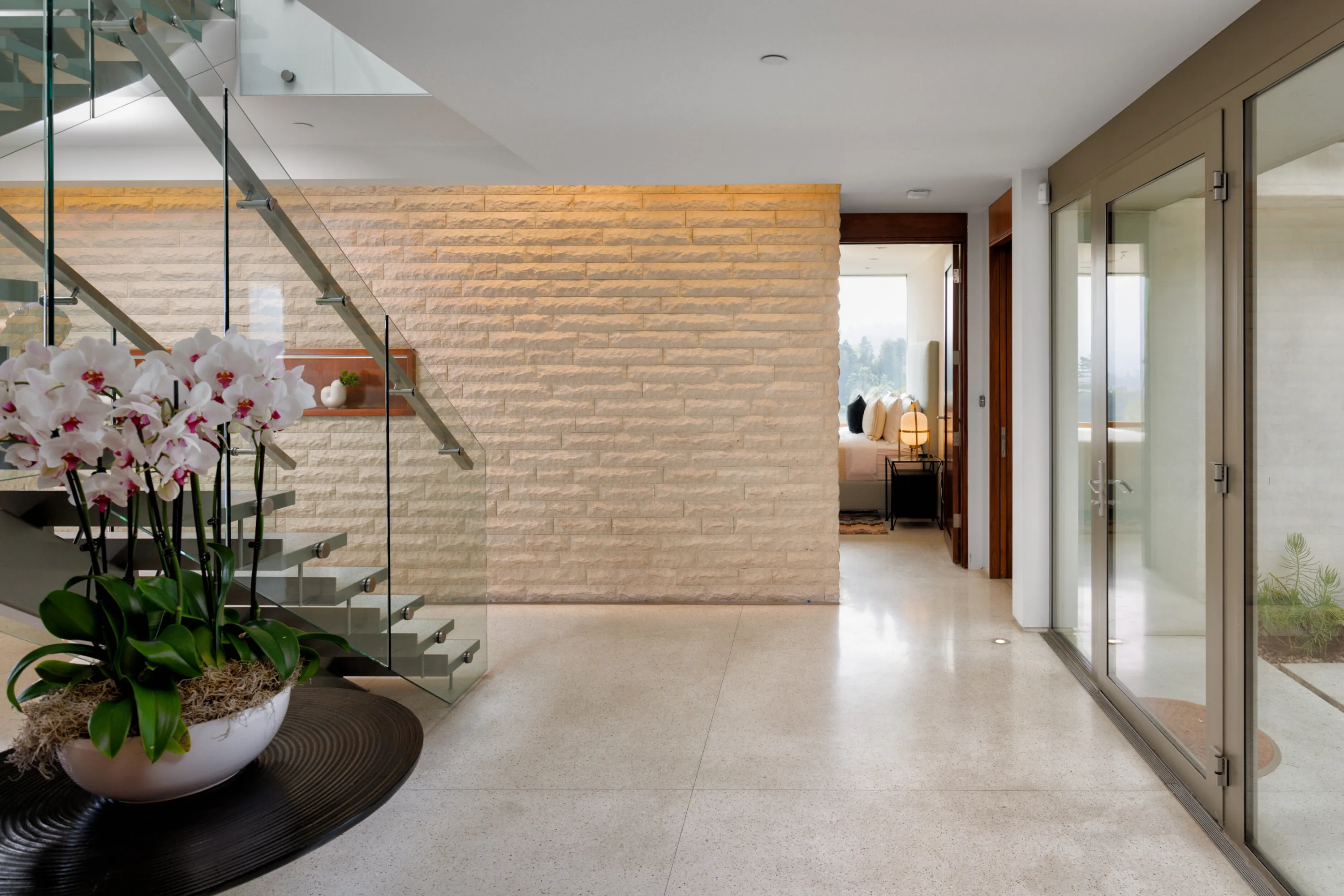 Entry hall with floating glass-railed staircase, backlit stone wall and orchids on a console
