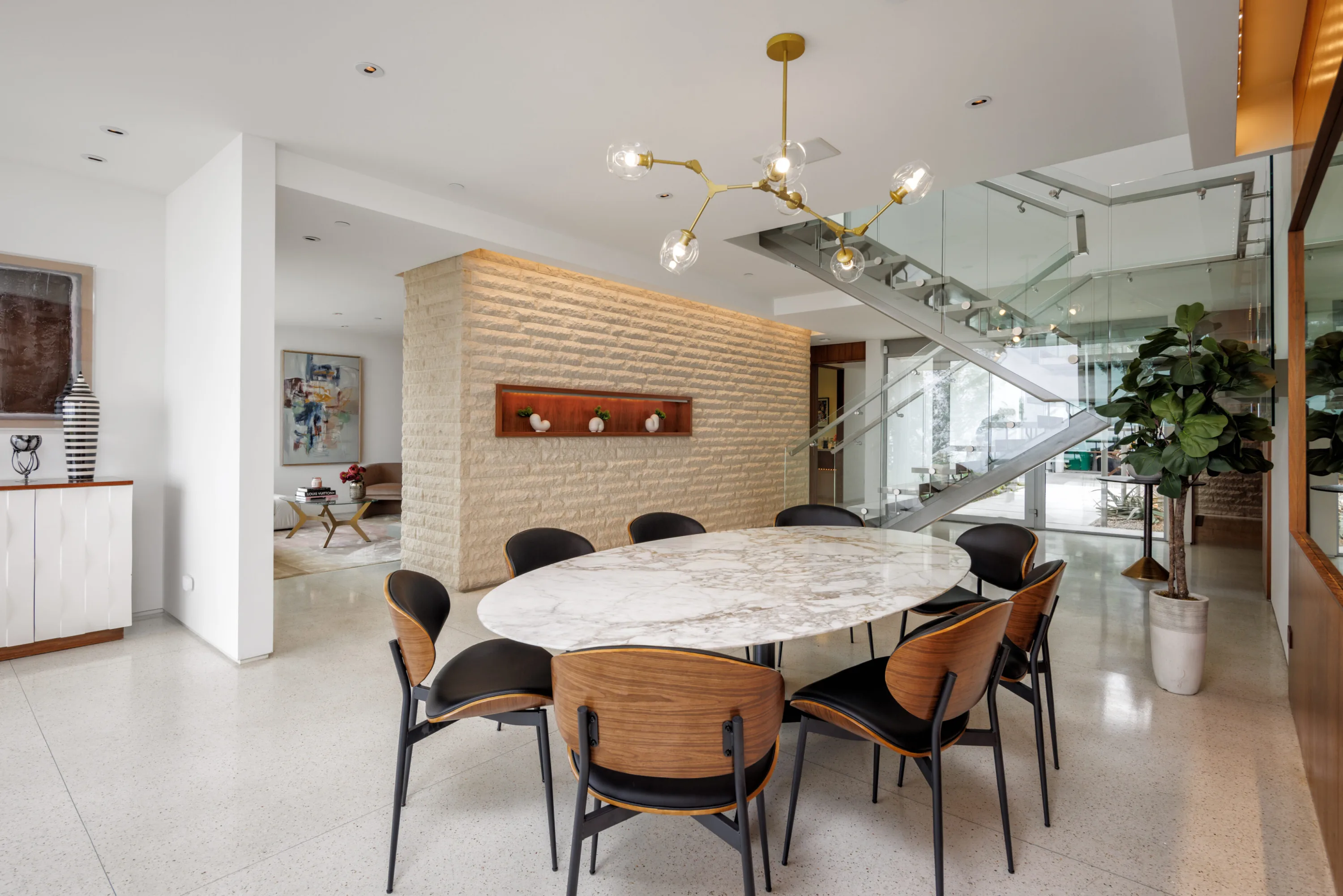 Oval marble dining table with walnut chairs under a brass globe chandelier beside the stone feature wall